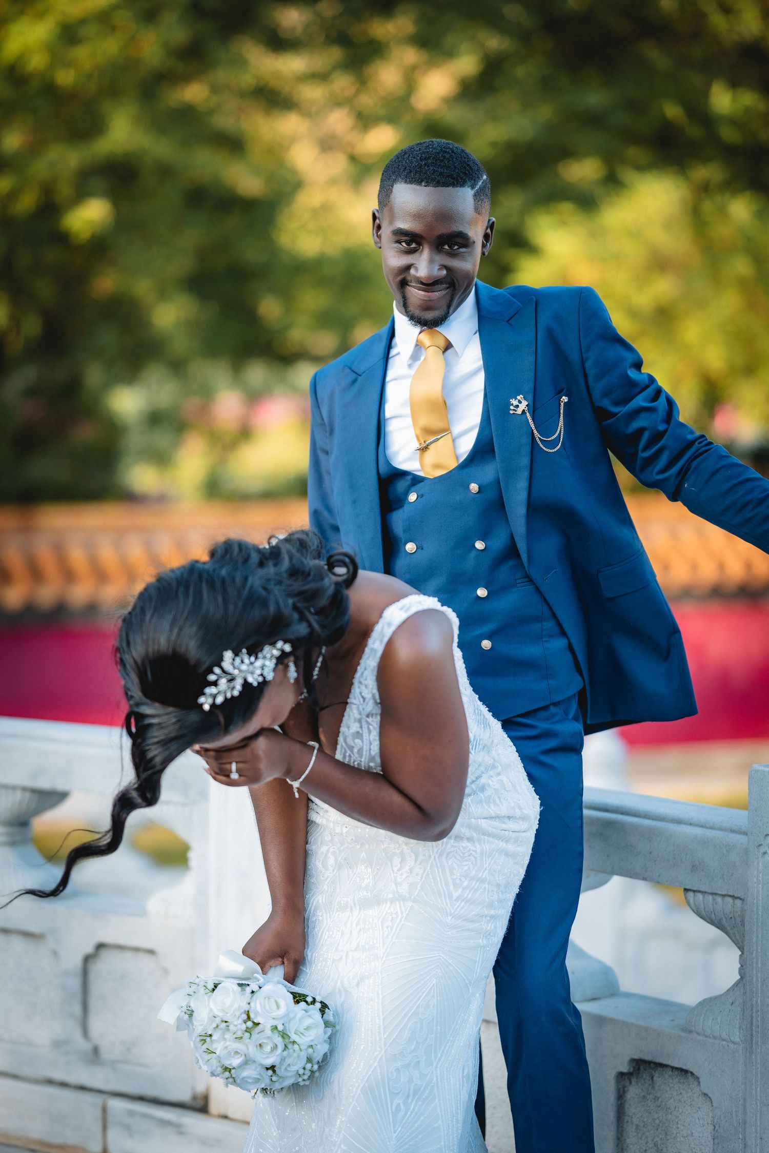 Groom in blue suit with gold tie poses behind bride in white lace dress holding white bouquet outdoors.
