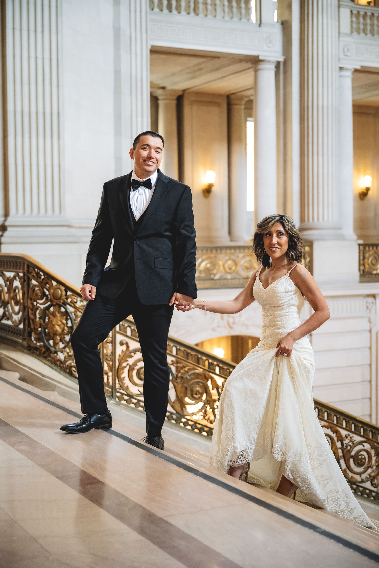 Elegant couple walking down ornate staircase at SF City Hall, groom in tuxedo leading bride in lace gown.