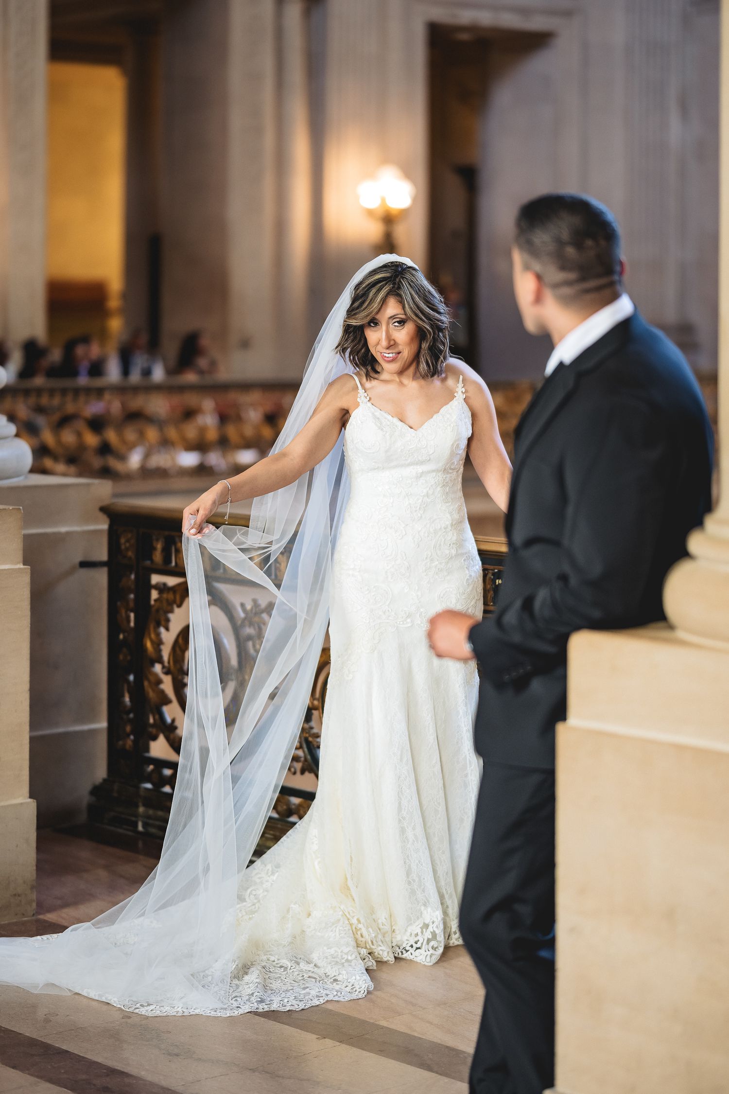 Bride in lace wedding gown and veil stands on steps facing groom in black tuxedo at elegant venue.