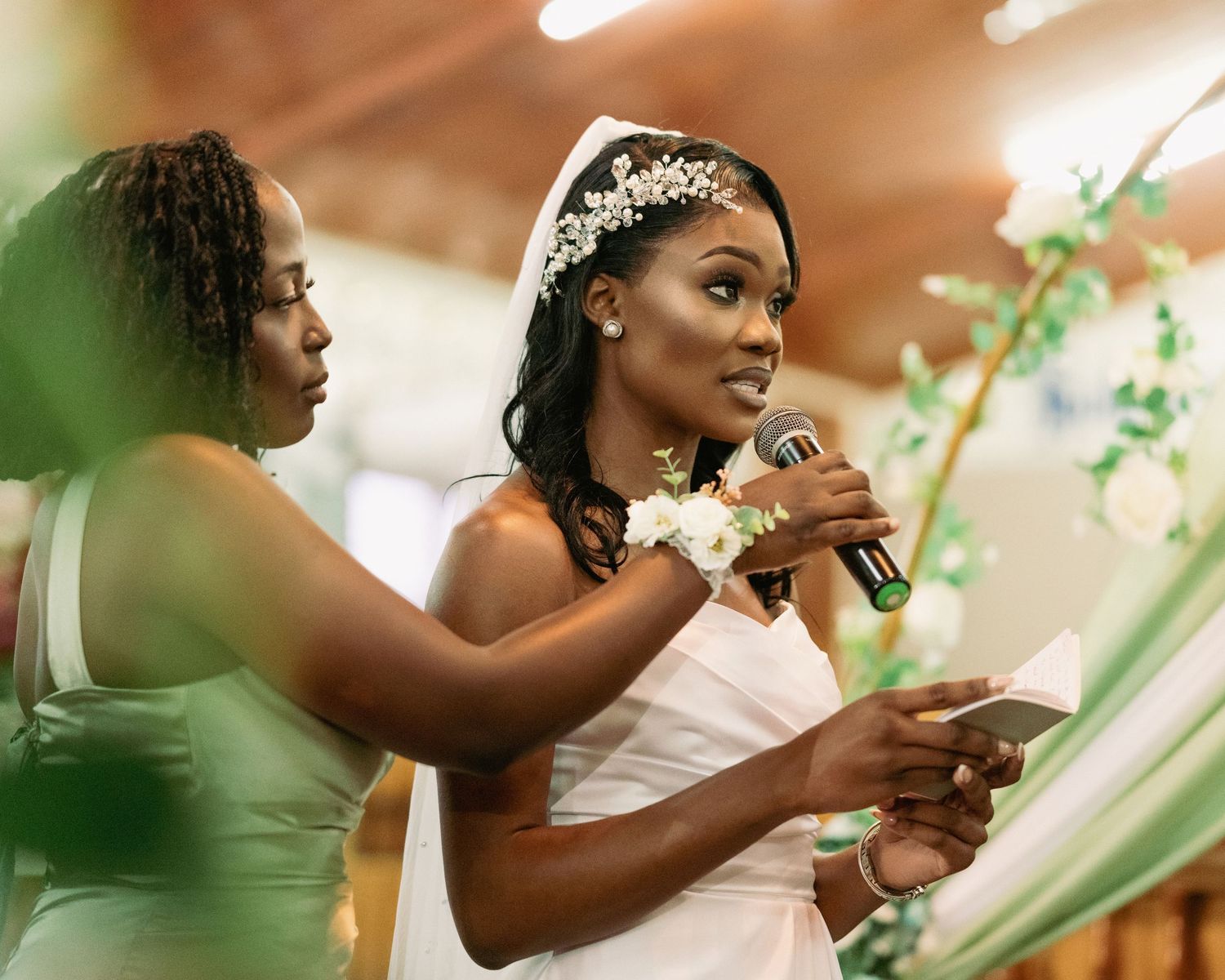 Beautiful Black bride in white gown reads vows into microphone assisted by bridesmaid in mint dress.