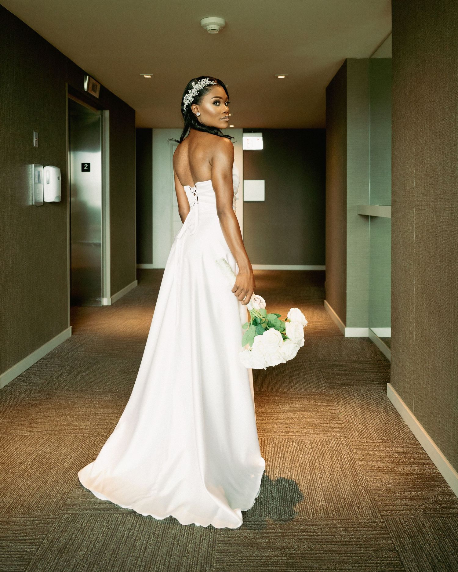 Elegant bride in white strapless gown holds bouquet while posing in hotel corridor, back to camera.