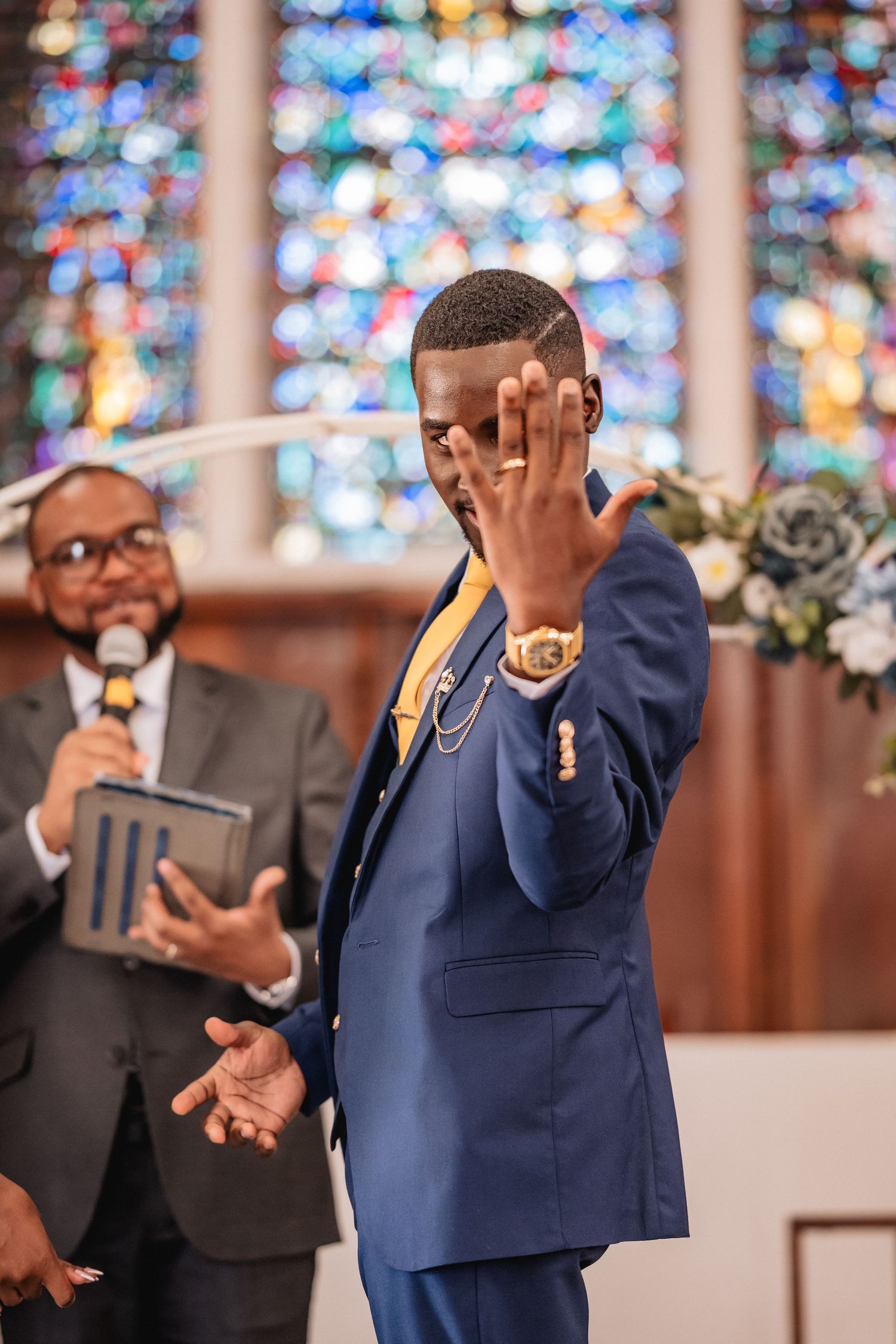 Groom in blue suit shows wedding ring at church altar with stained glass windows and officiant in background.