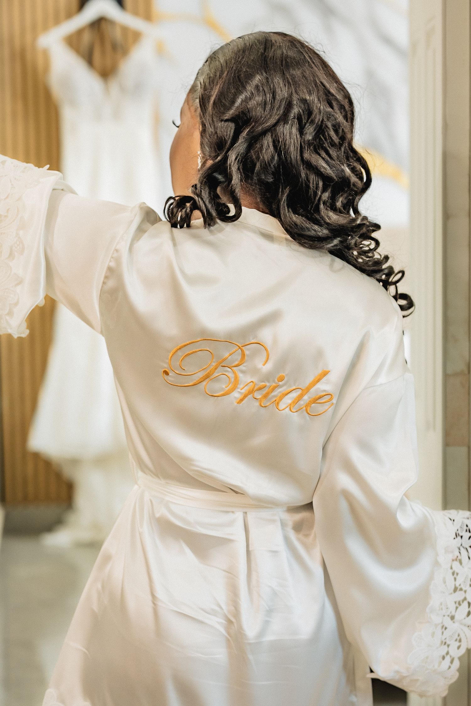 Bride in white satin robe with gold embroidered text admires wedding dress in bridal boutique.