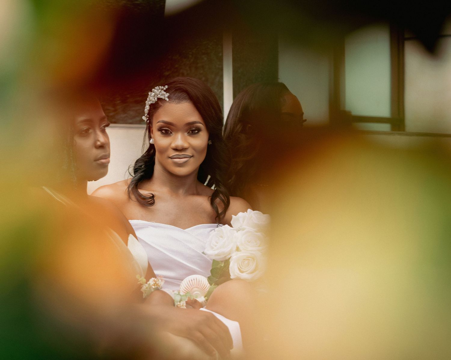 Beautiful Black bride in white off-shoulder gown holding white roses, photographed through colorful bokeh foreground.