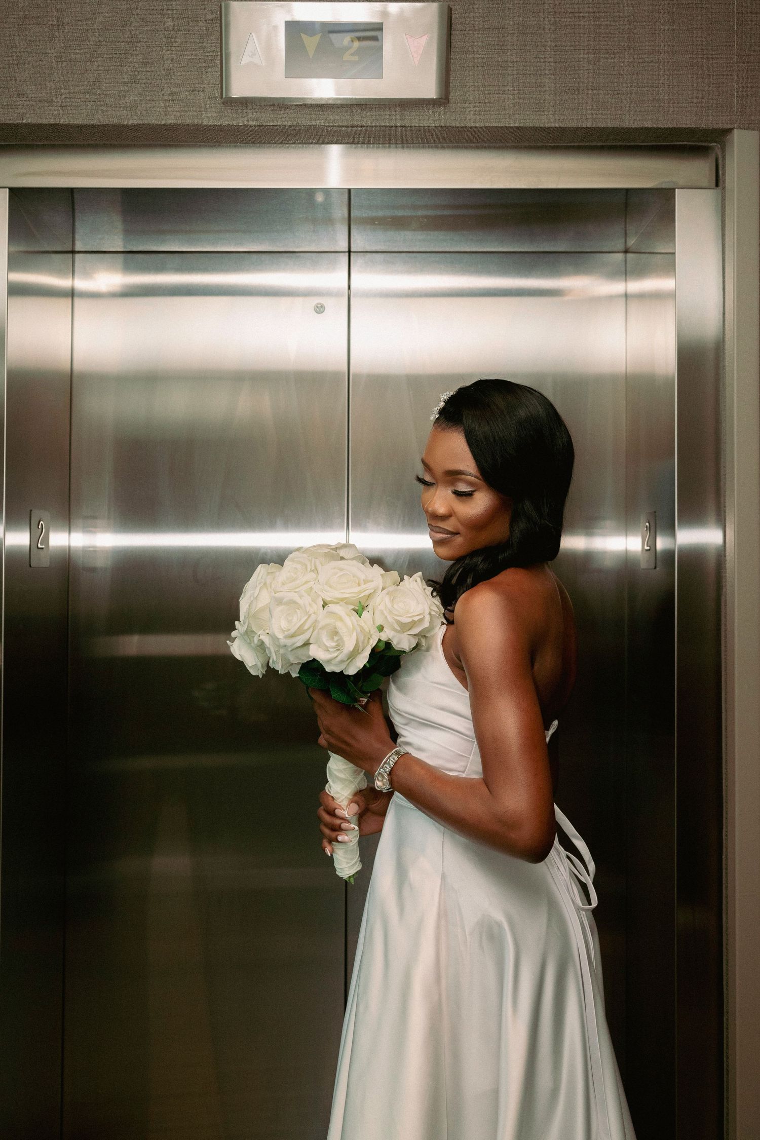Elegant bride in white satin gown holding white rose bouquet standing in front of silver elevator doors.