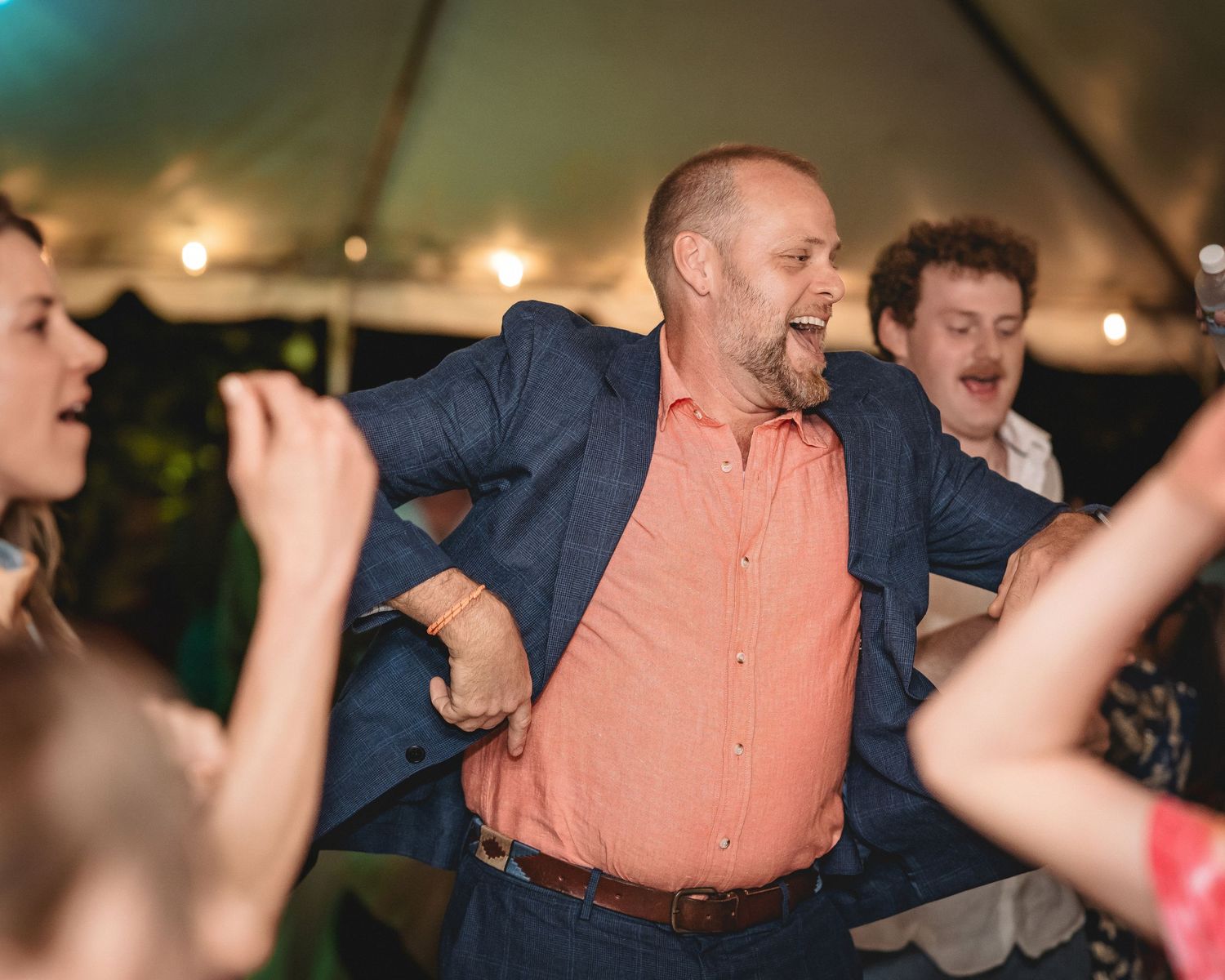 Man in coral shirt and blue blazer dancing energetically at a festive tent party with other guests.