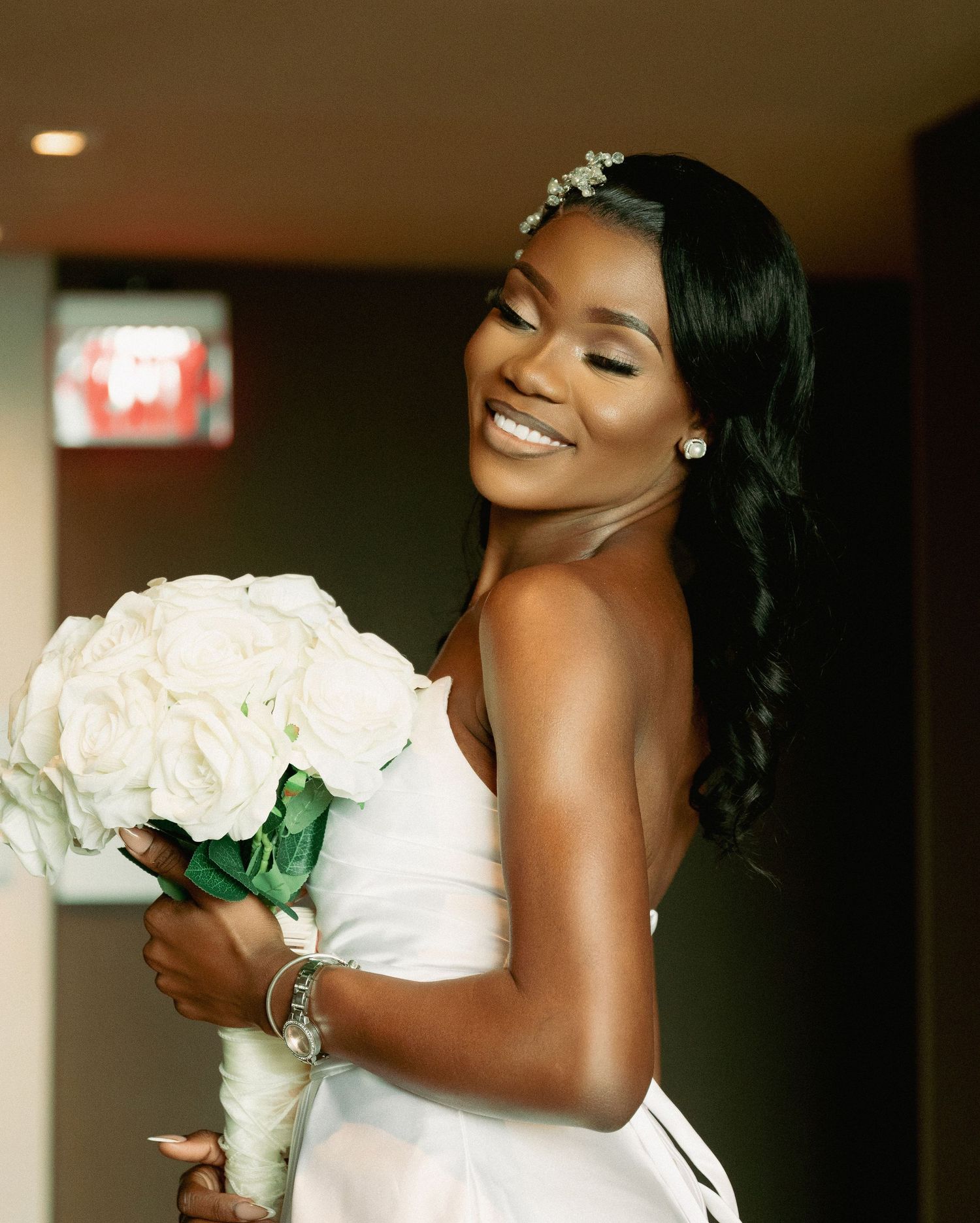 Radiant Black bride smiling joyfully in white strapless dress holding white rose bouquet with pearl earrings.