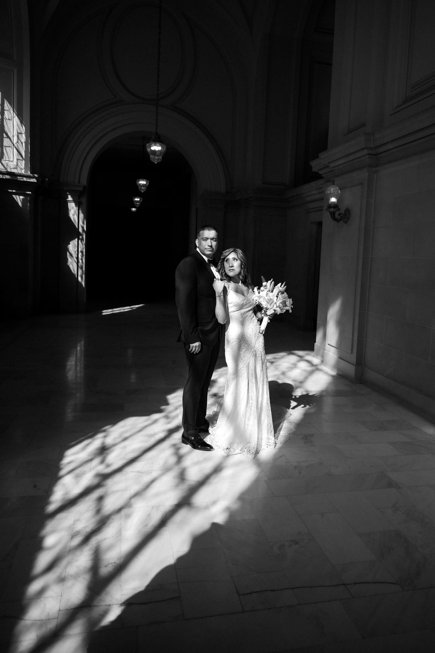 Black and white wedding photo of bride and groom standing in dramatic light inside an elegant hall.