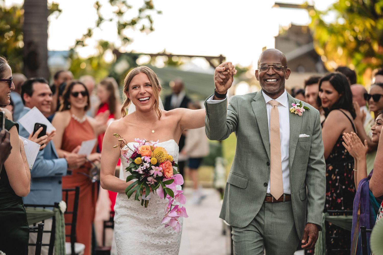 Joyful interracial couple walks down wedding aisle, bride holding colorful bouquet, groom in sage suit, guests cheering outdoors.
