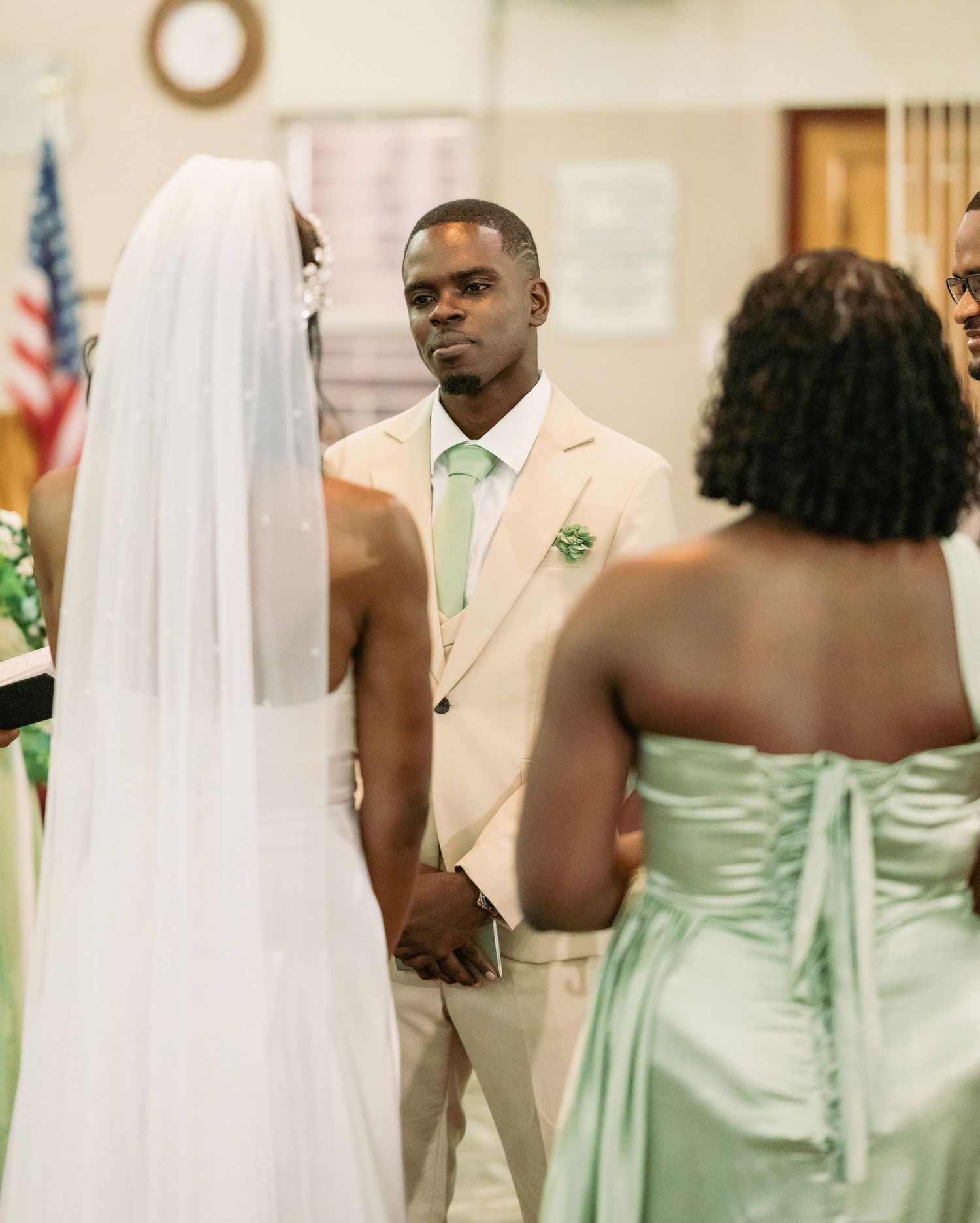 Groom in beige suit with mint tie gazes at bride during church wedding ceremony at altar.
