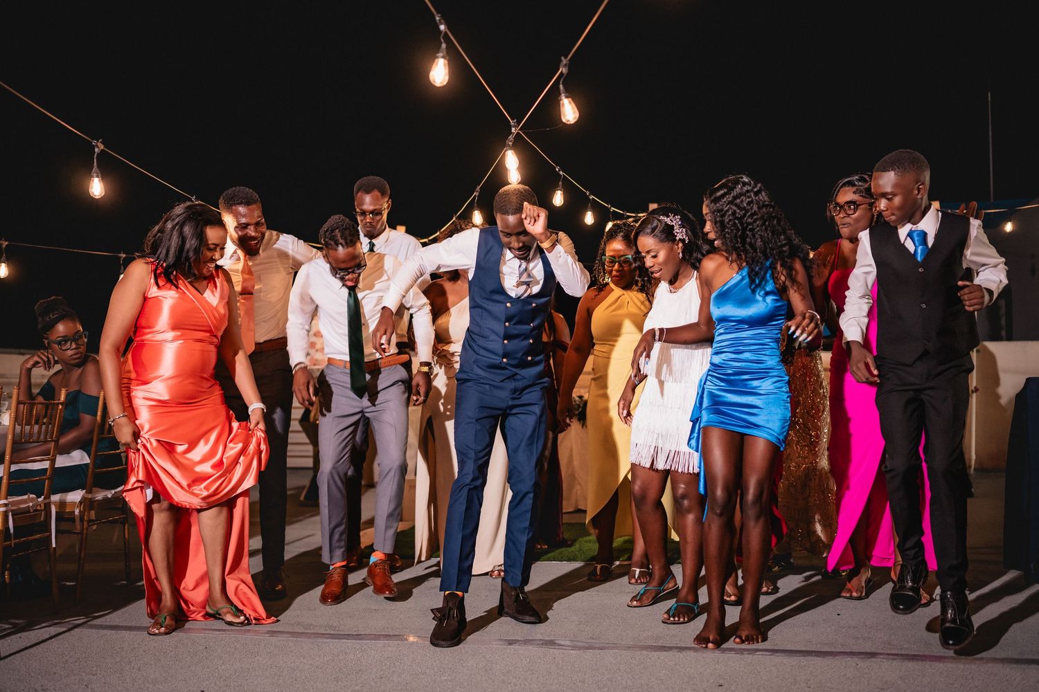 Wedding guests dance joyfully under string lights at an outdoor night reception in colorful formal attire.