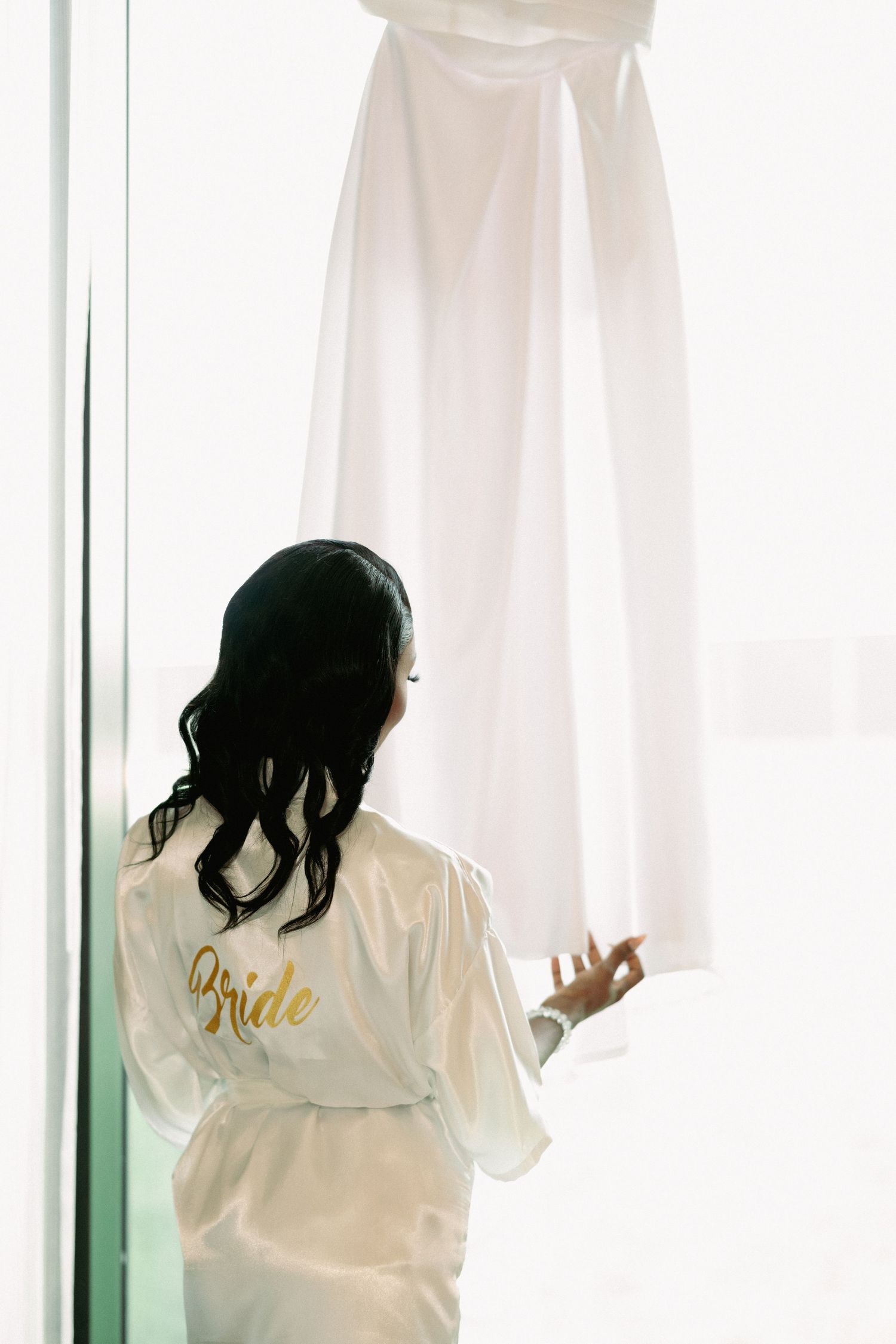 Bride in white satin robe with gold script stands before hanging wedding dress in bright window light.
