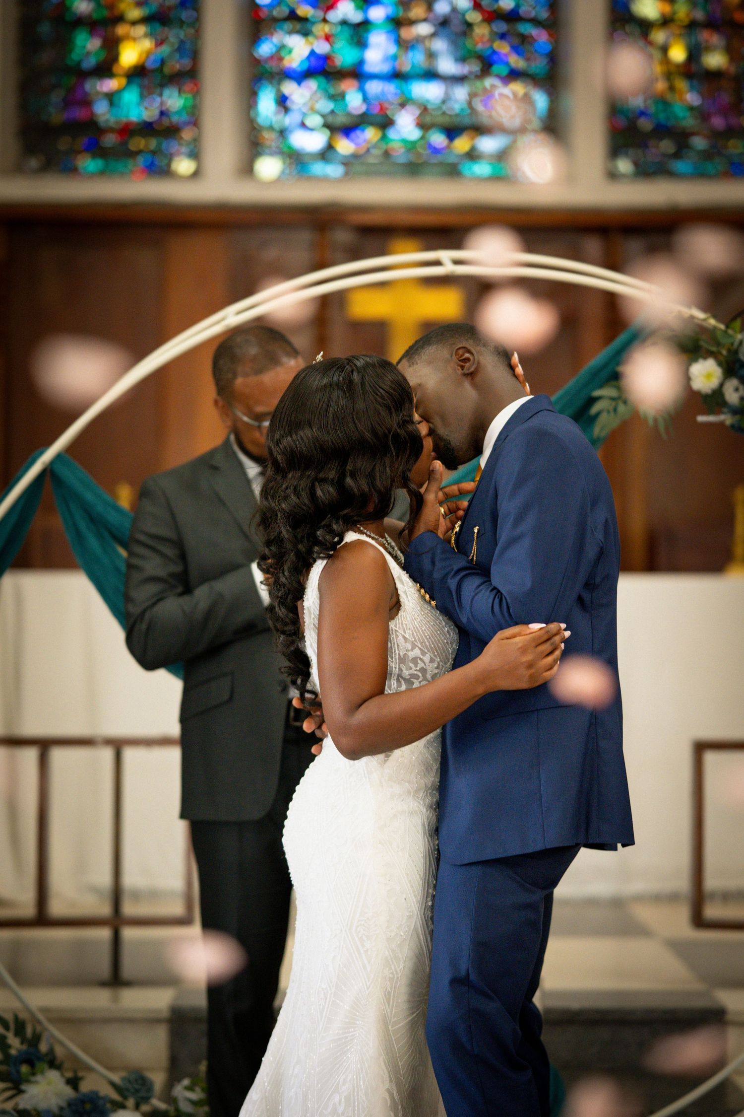 Bride and groom share first kiss at church altar under circular arch with stained glass backdrop.