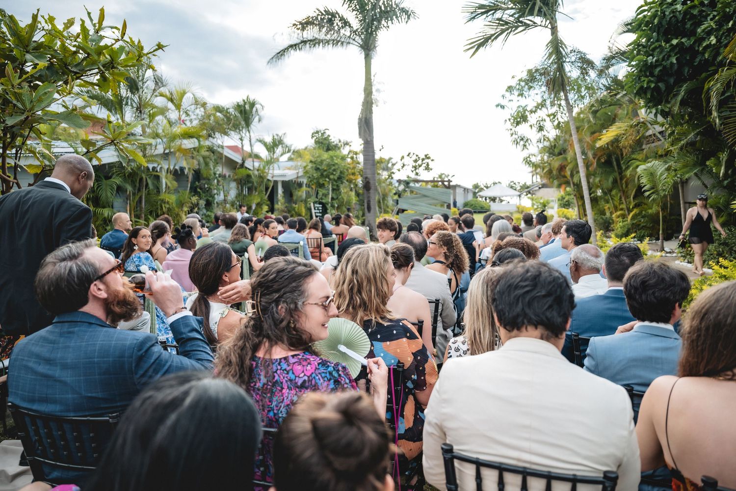 Outdoor wedding ceremony with guests seated in chairs surrounded by lush tropical palm trees and greenery.