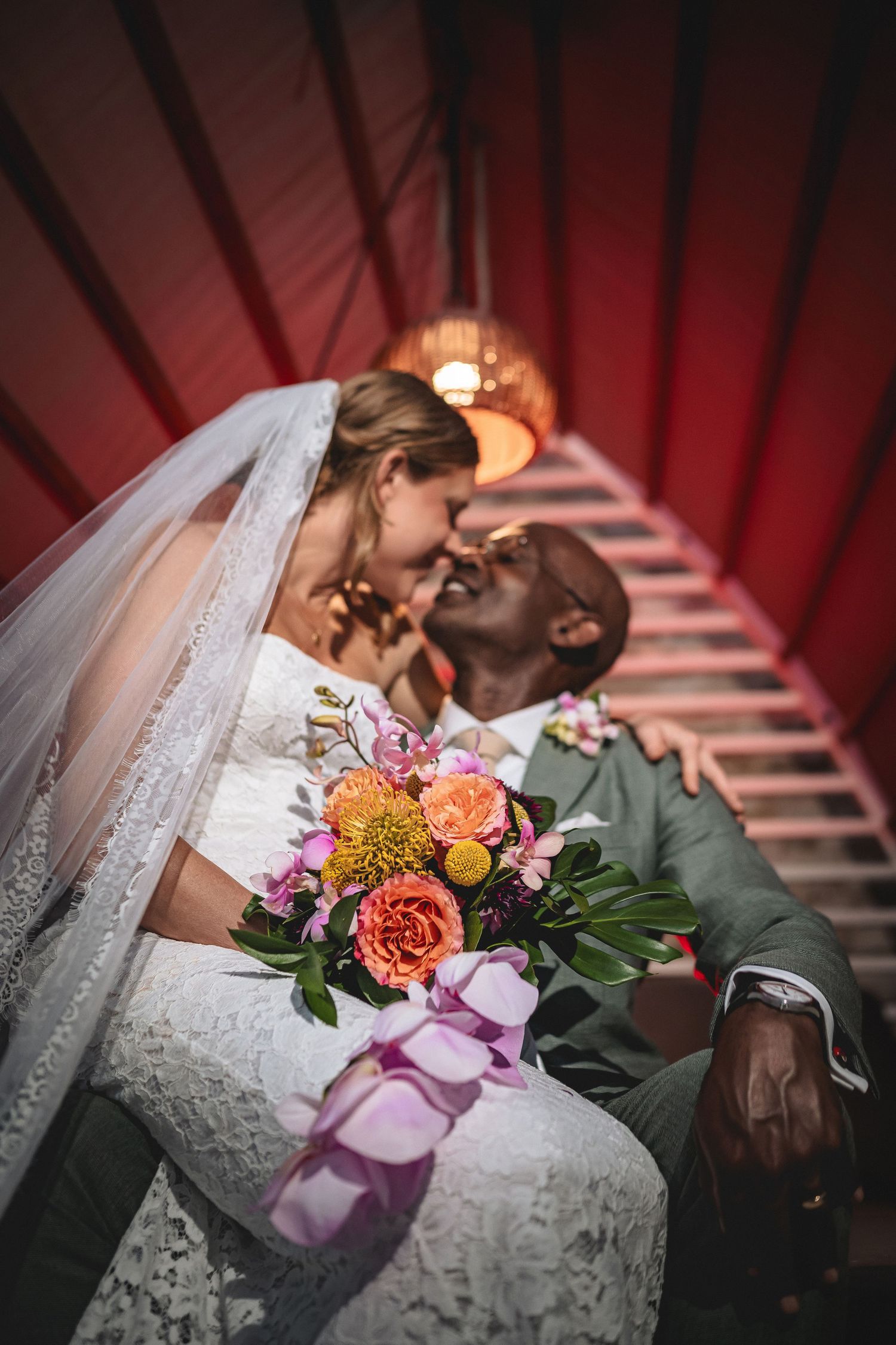 Interracial couple sharing a romantic kiss on their wedding day, bride holding a colorful bouquet of orange and pink flowers.