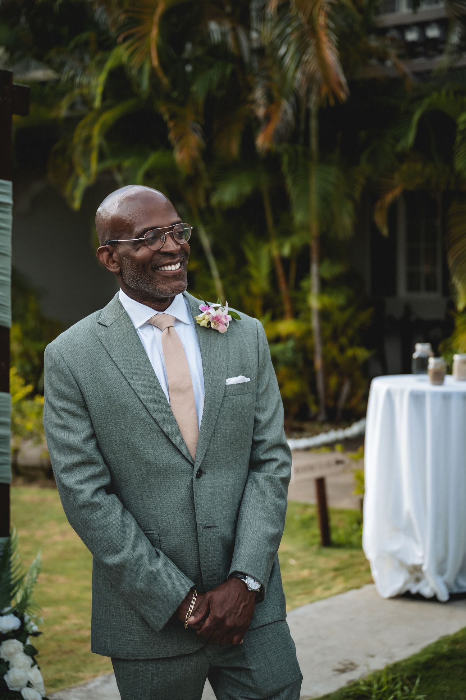 Smiling groom in gray suit with boutonniere stands outdoors at tropical wedding ceremony.