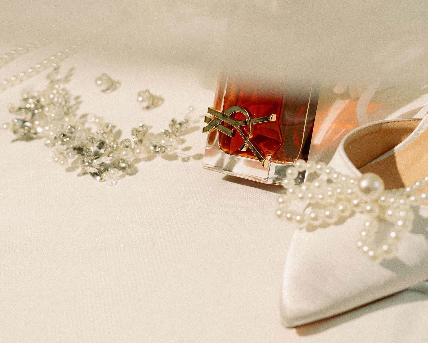 Elegant bridal accessories including pearl heels, crystal hair piece, and luxury perfume bottle on white satin.