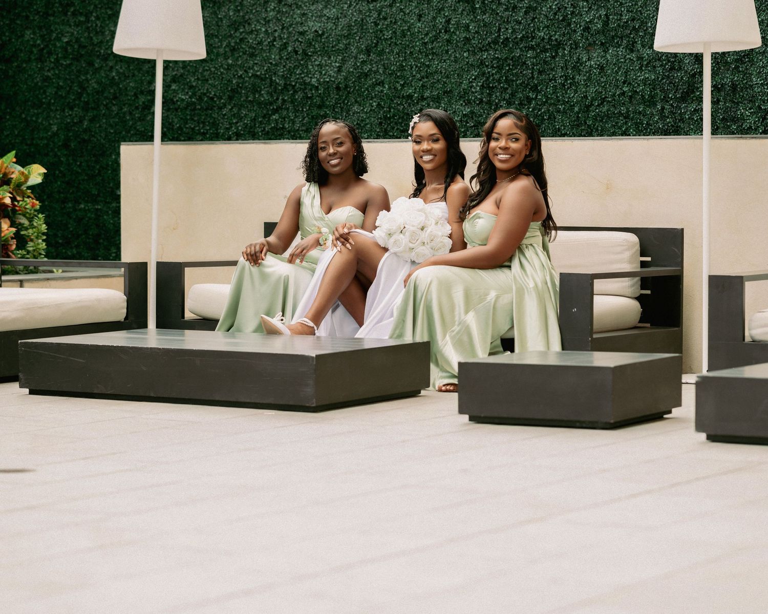A bride in white sits with two bridesmaids in sage green gowns outdoors.