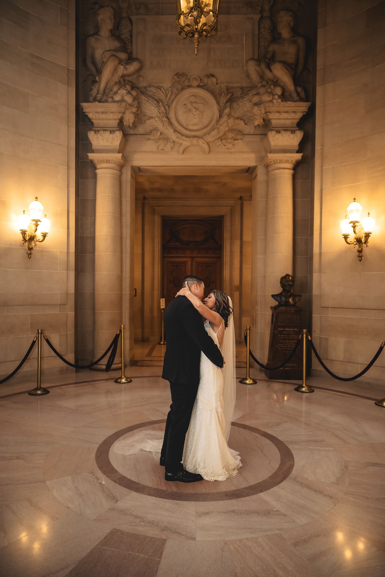 Bride and groom share a romantic kiss in an elegant marble rotunda with ornate sculptures and warm golden sconce lighting.