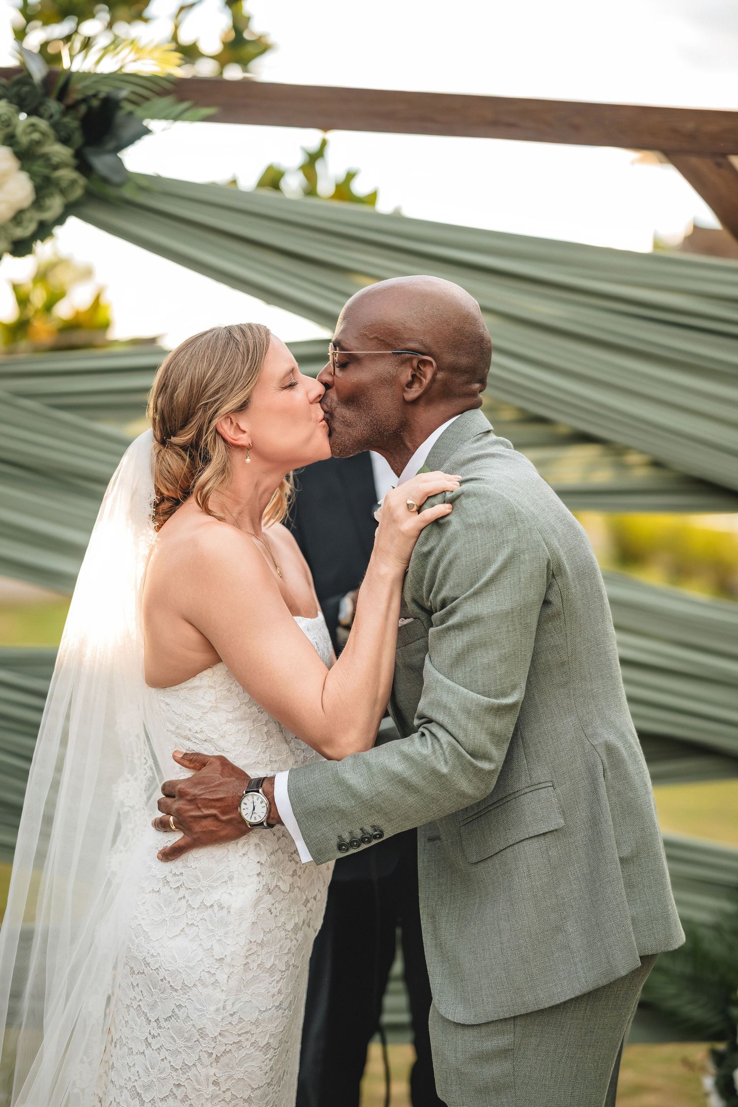 Interracial couple sharing their first kiss as newlyweds at an outdoor wedding ceremony with elegant green draping.