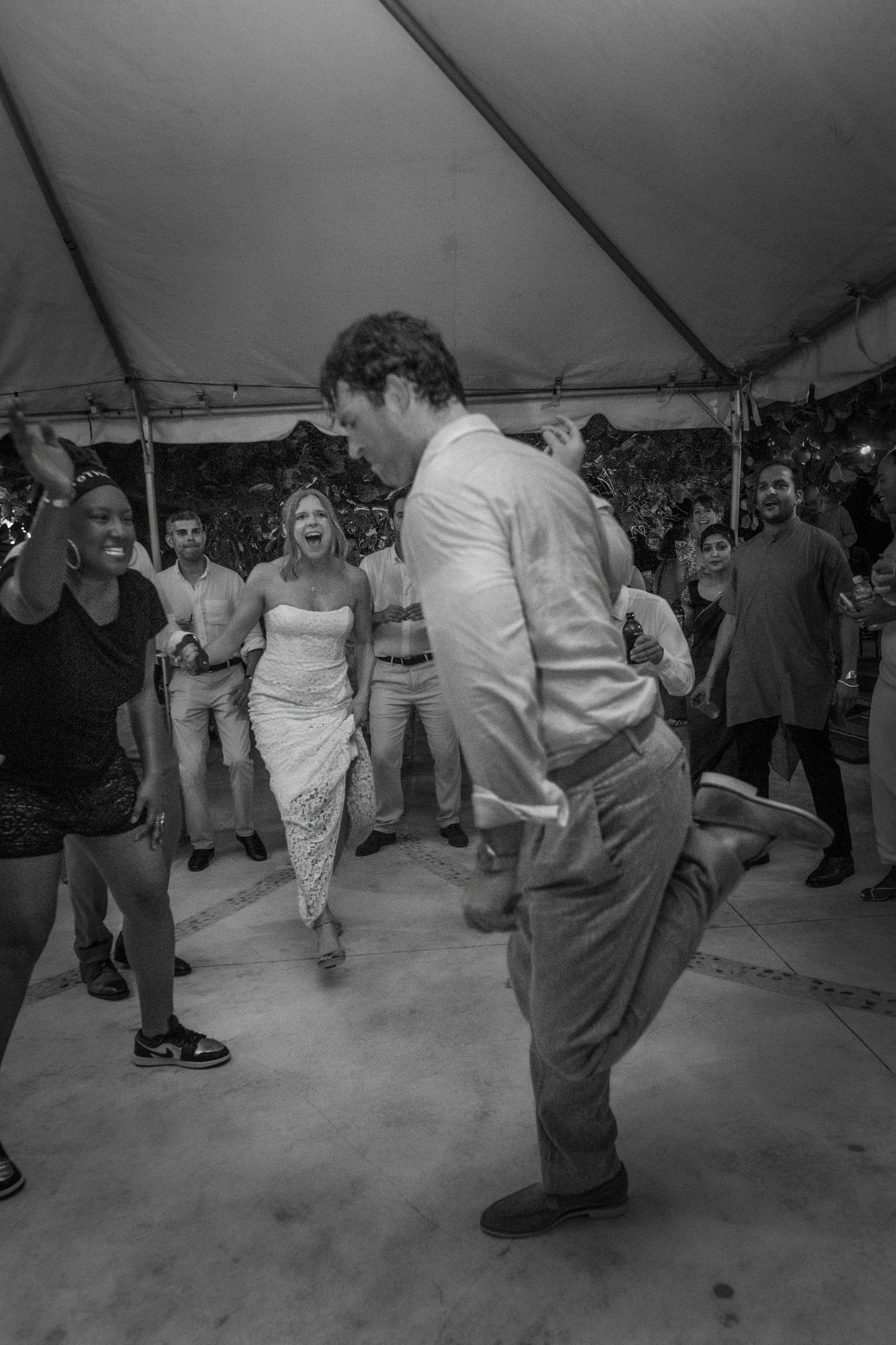 Black and white photo of a bride and groom dancing joyfully under a tent surrounded by cheering wedding guests.