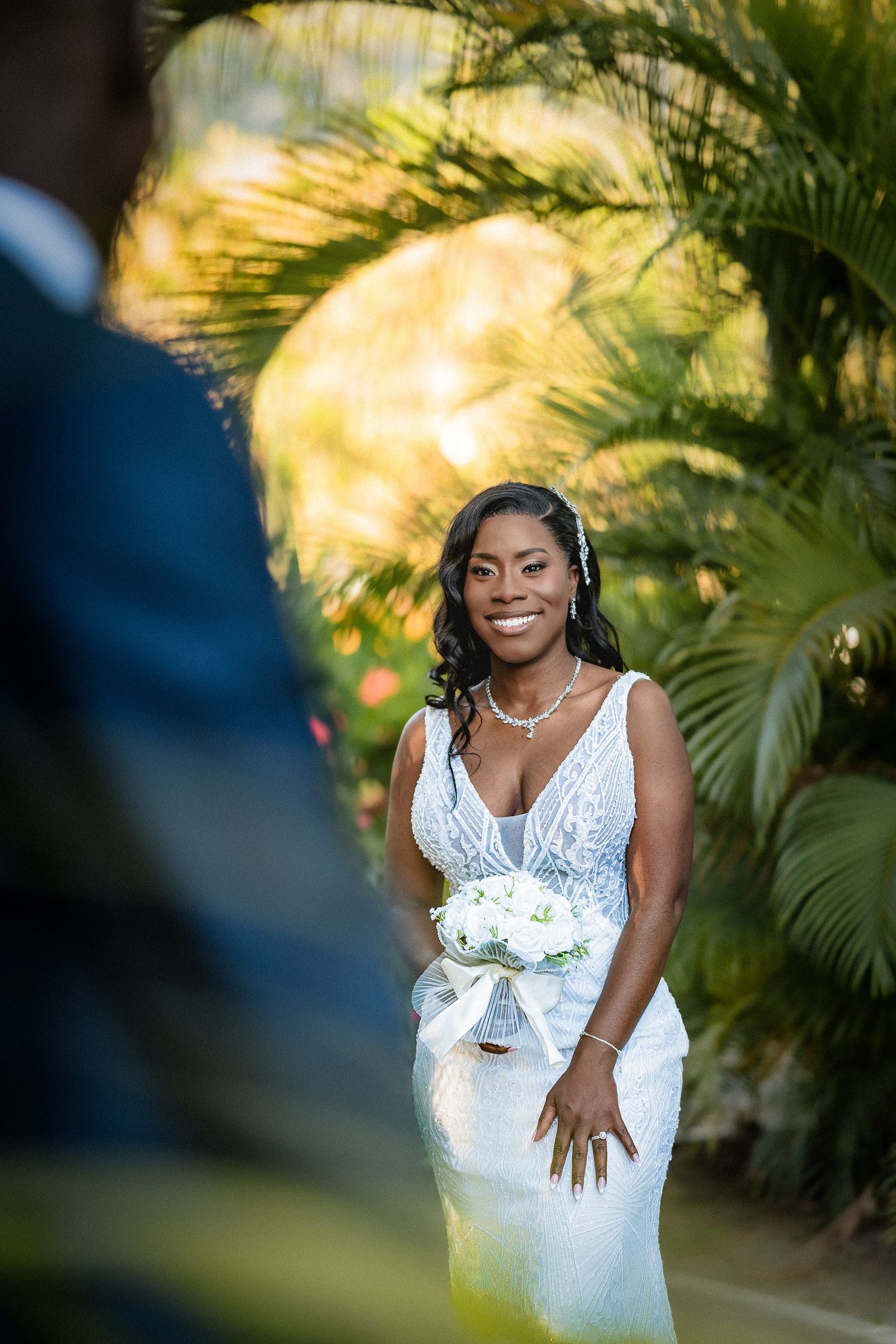 Beautiful Black bride holding white bouquet in fitted lace wedding dress among tropical palm trees.