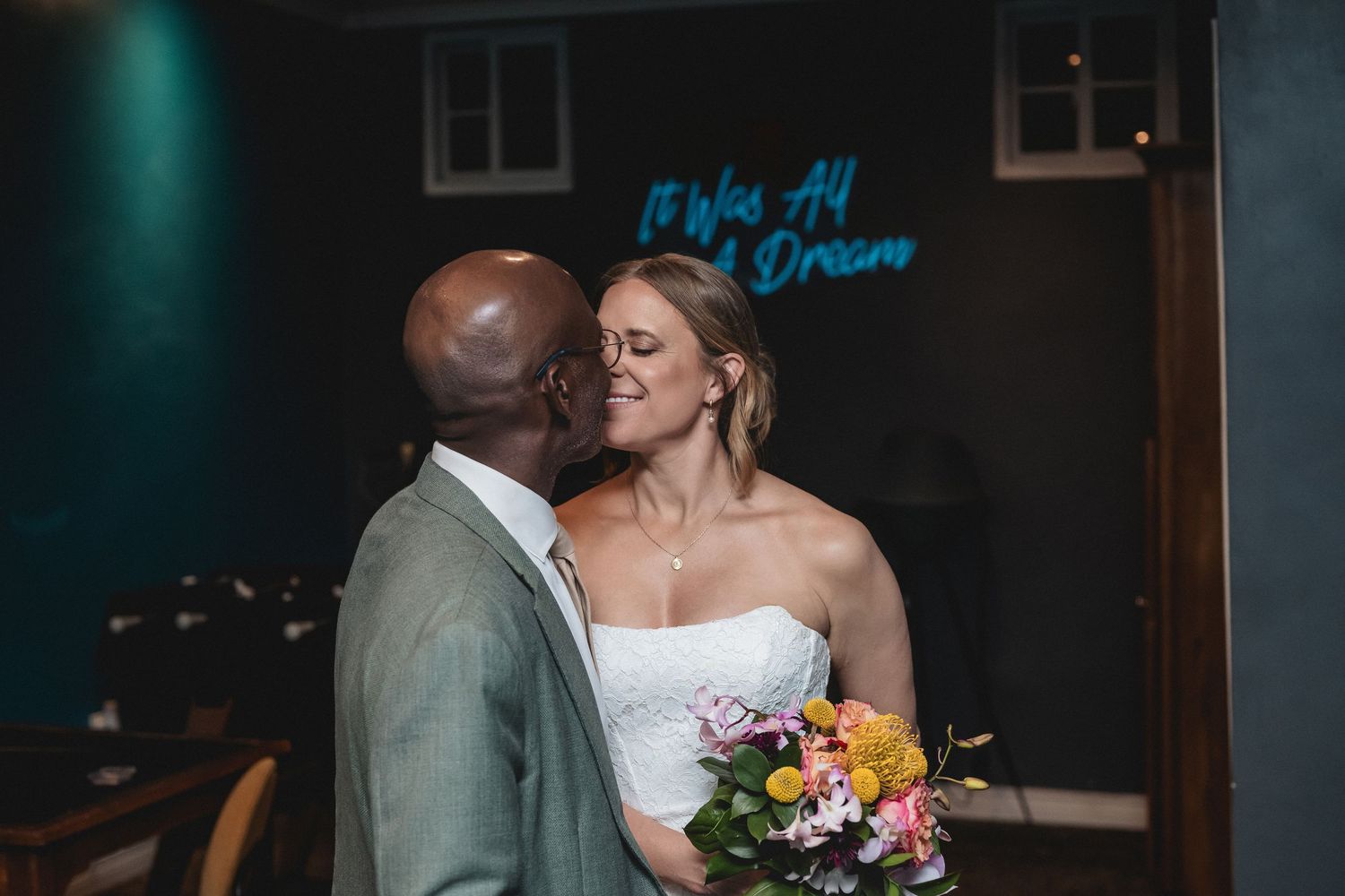 A newlywed couple shares a romantic moment, bride holding colorful bouquet, with a blue neon sign in the background.