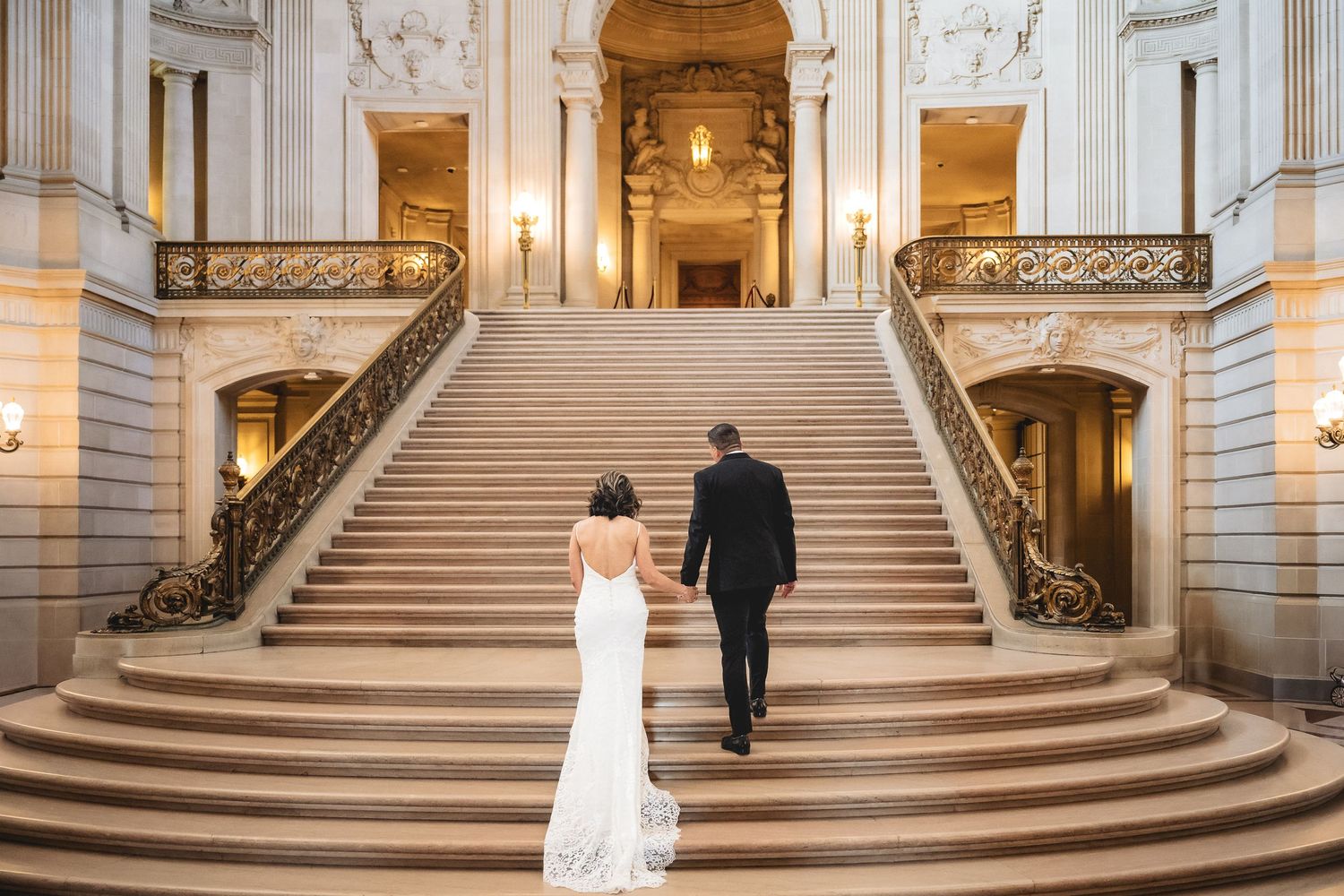 Bride and groom walking hand in hand up grand staircase at San Francisco City Hall wedding venue.