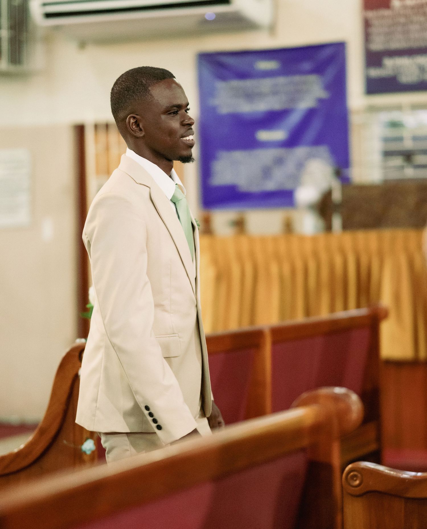 Young man in beige suit with green tie standing in church pews during a religious service.
