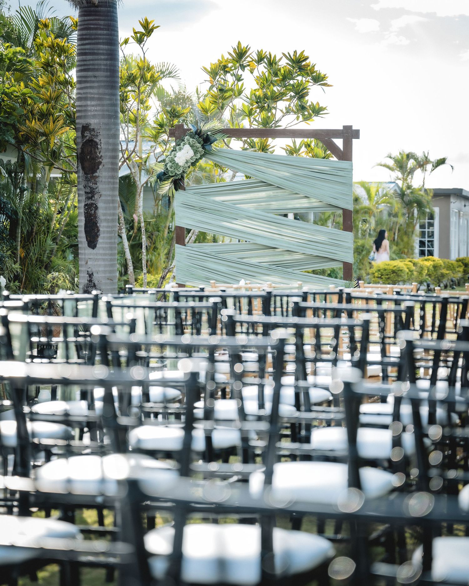 Outdoor wedding ceremony setup with clear chiavari chairs and a white ribbon-draped wooden arch surrounded by tropical greenery.