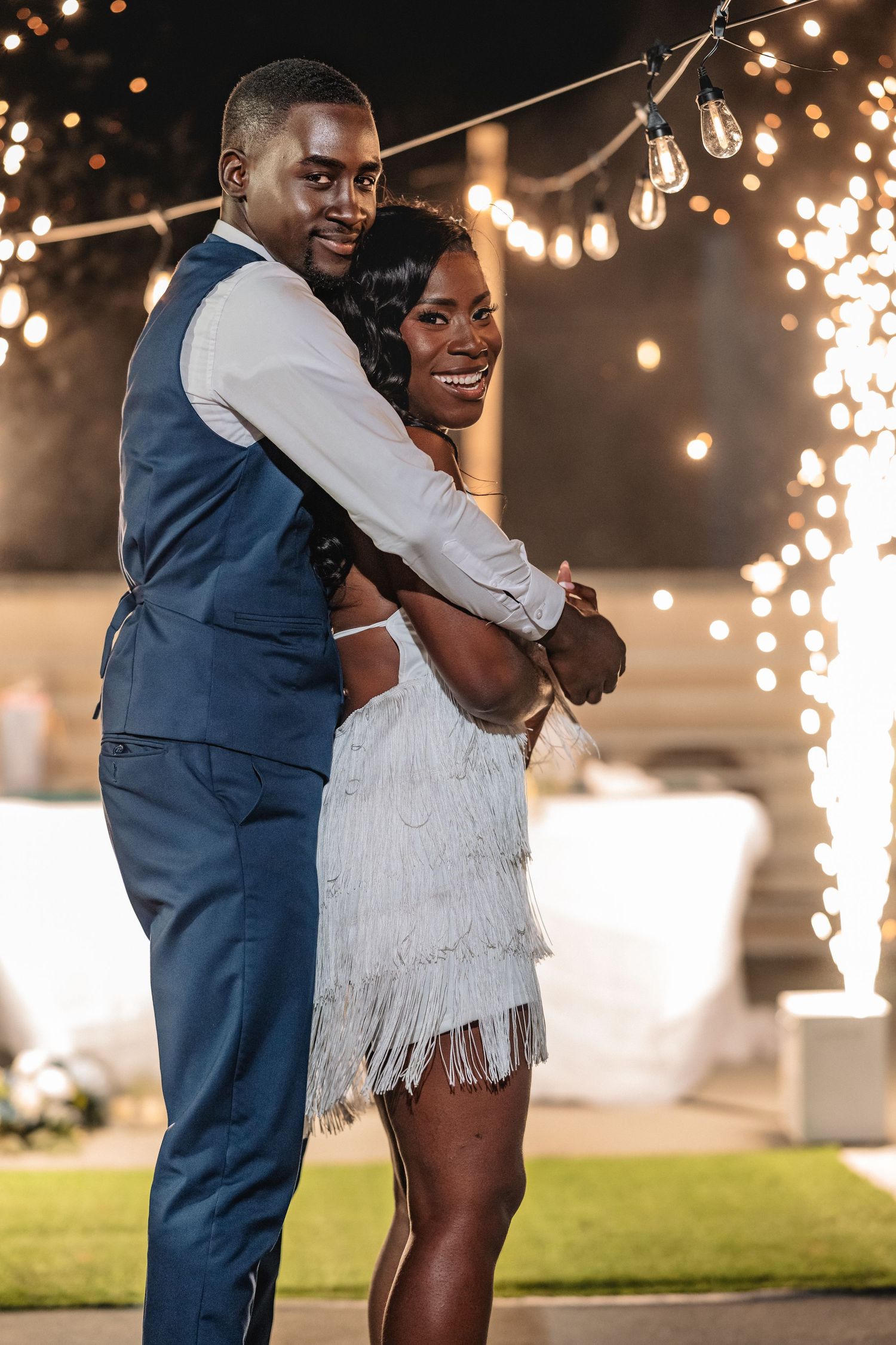 Smiling newlyweds embrace during first dance with sparkling lights at outdoor evening reception.