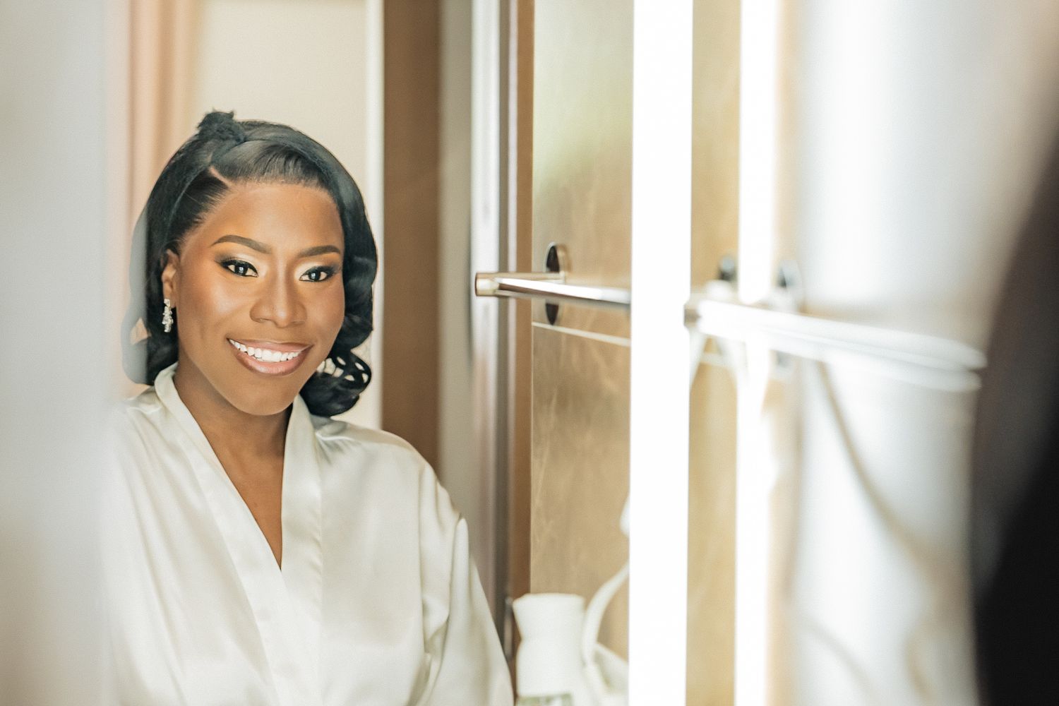 Smiling Black bride in white robe getting ready, reflected in a lighted bathroom mirror.