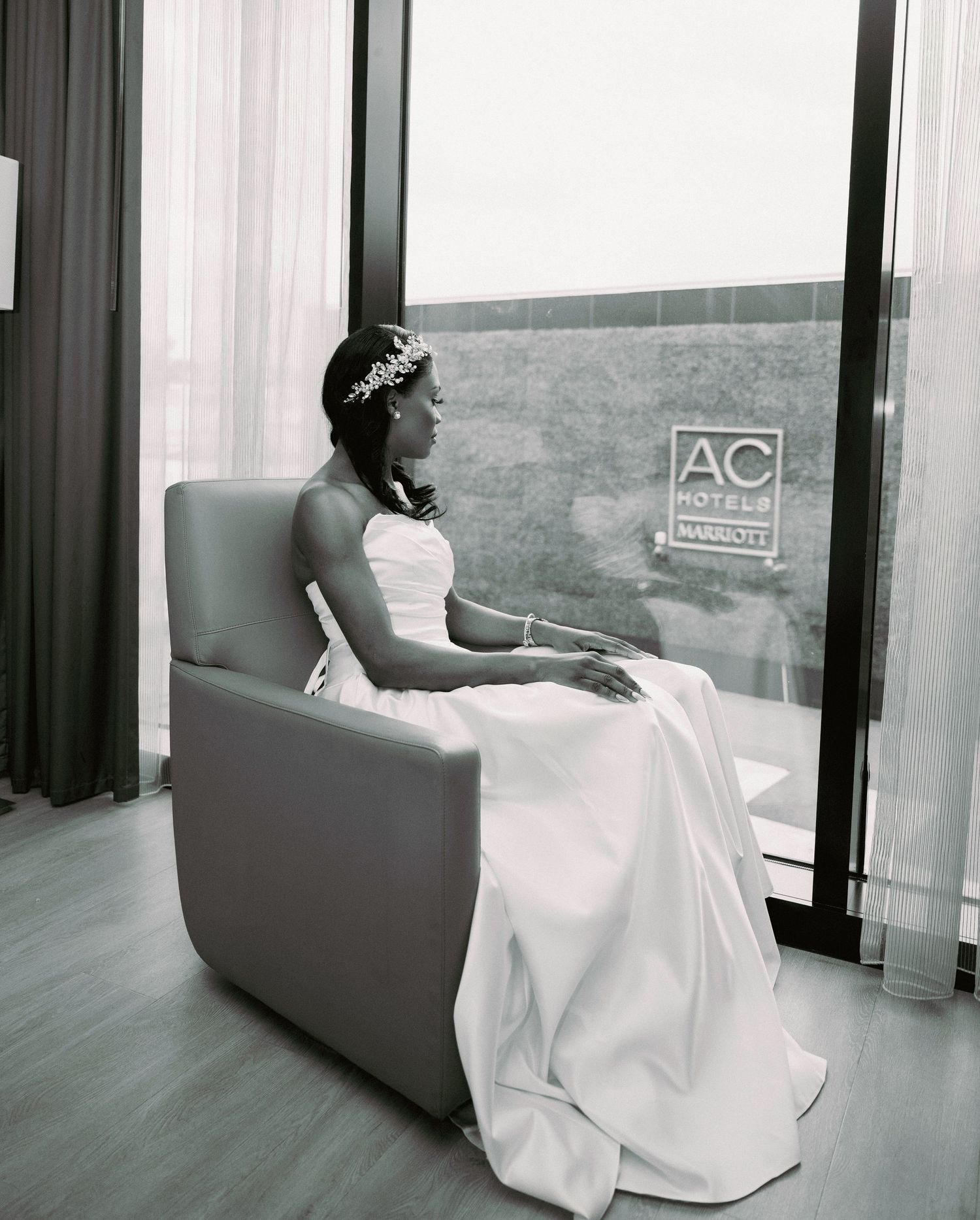 Bride in white gown sitting in chair by window at AC Hotels Marriott in black and white photo.