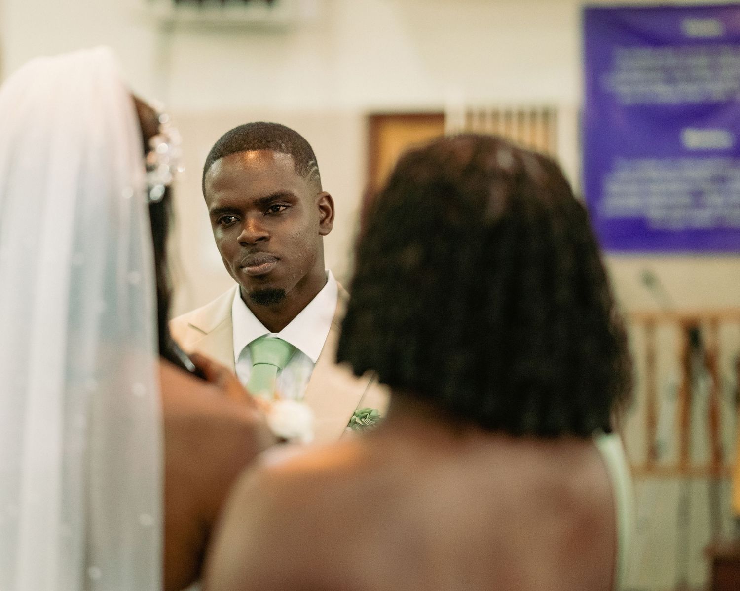 Groom in a beige suit with green tie gazing at his bride during a church wedding ceremony.