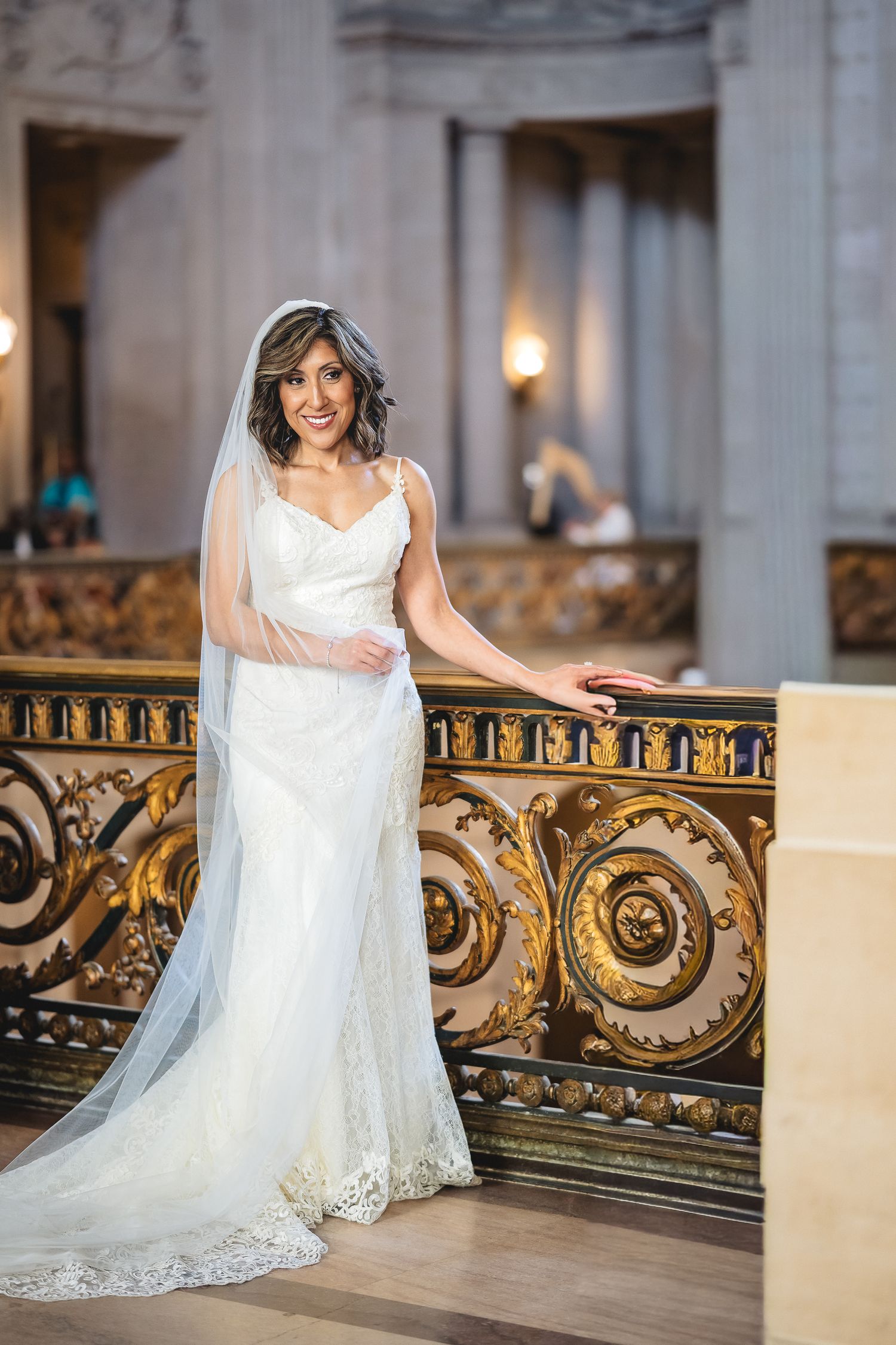 Bride in white lace gown and veil poses gracefully by ornate gold railing inside elegant wedding venue.