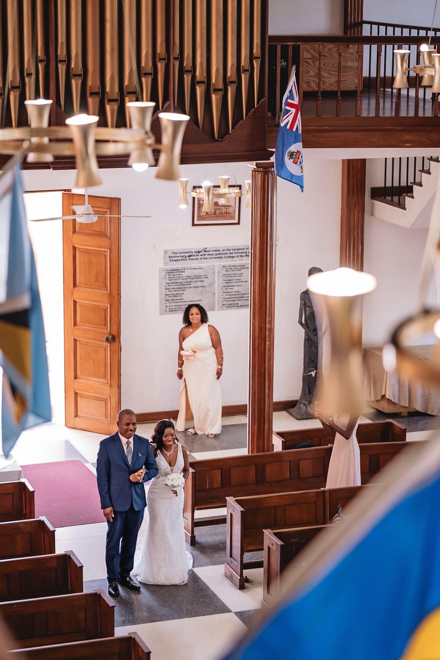 Aerial view of bride walking down church aisle with escort, Cayman Islands flags visible, organ pipes above.
