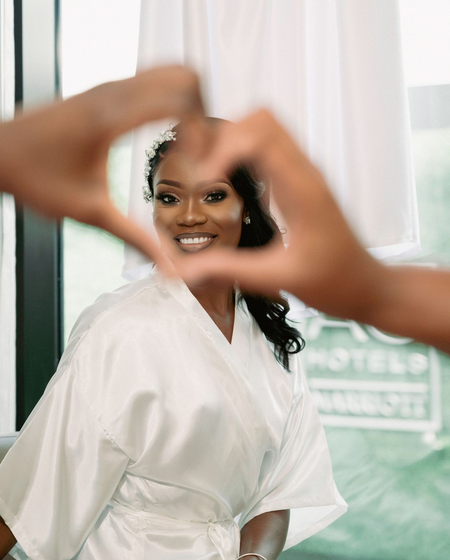 Smiling bride in white satin robe with floral hair accessory, framed by heart-shaped hands gesture.