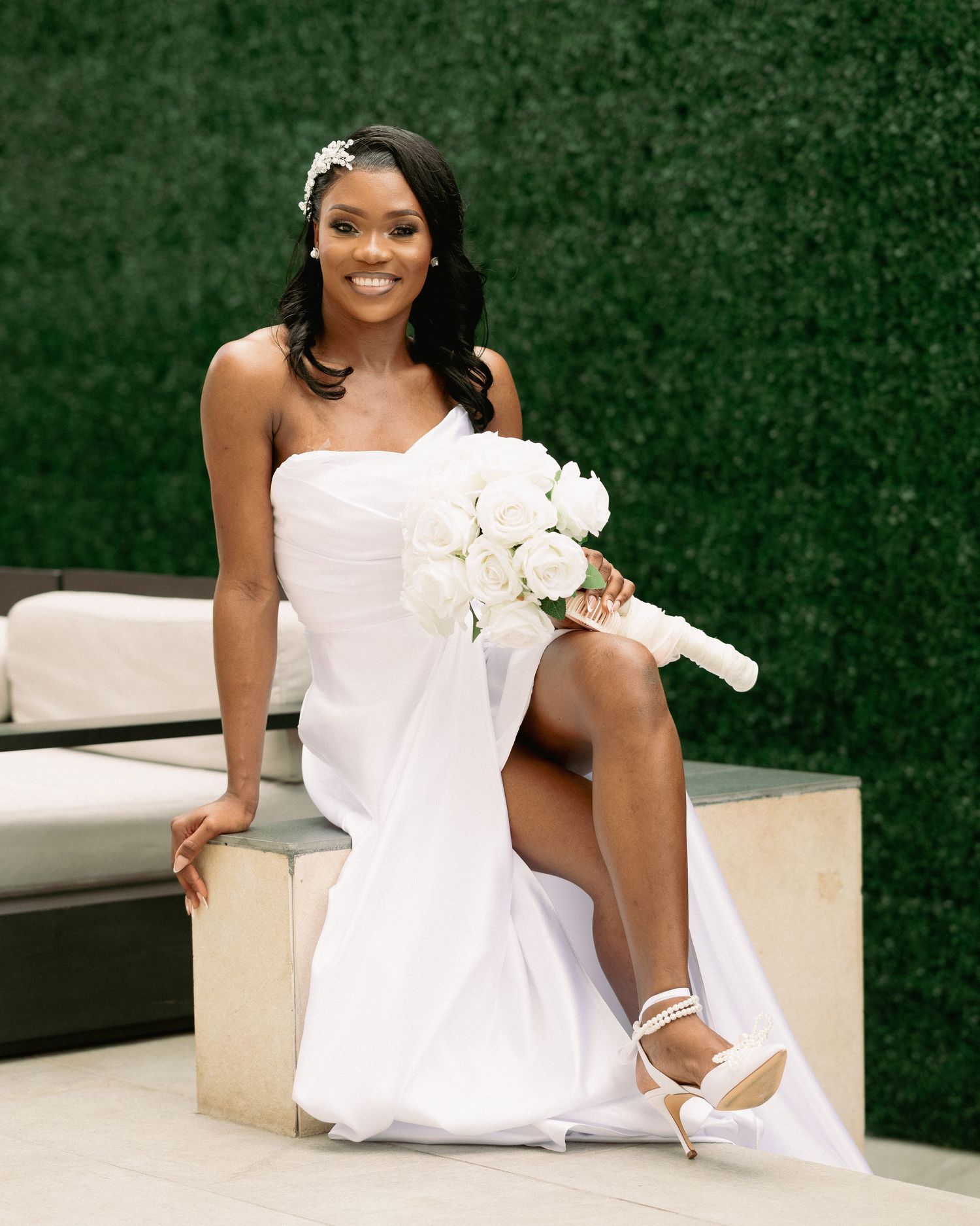 Smiling bride in white strapless gown with rose bouquet seated outdoors against green hedge wall.