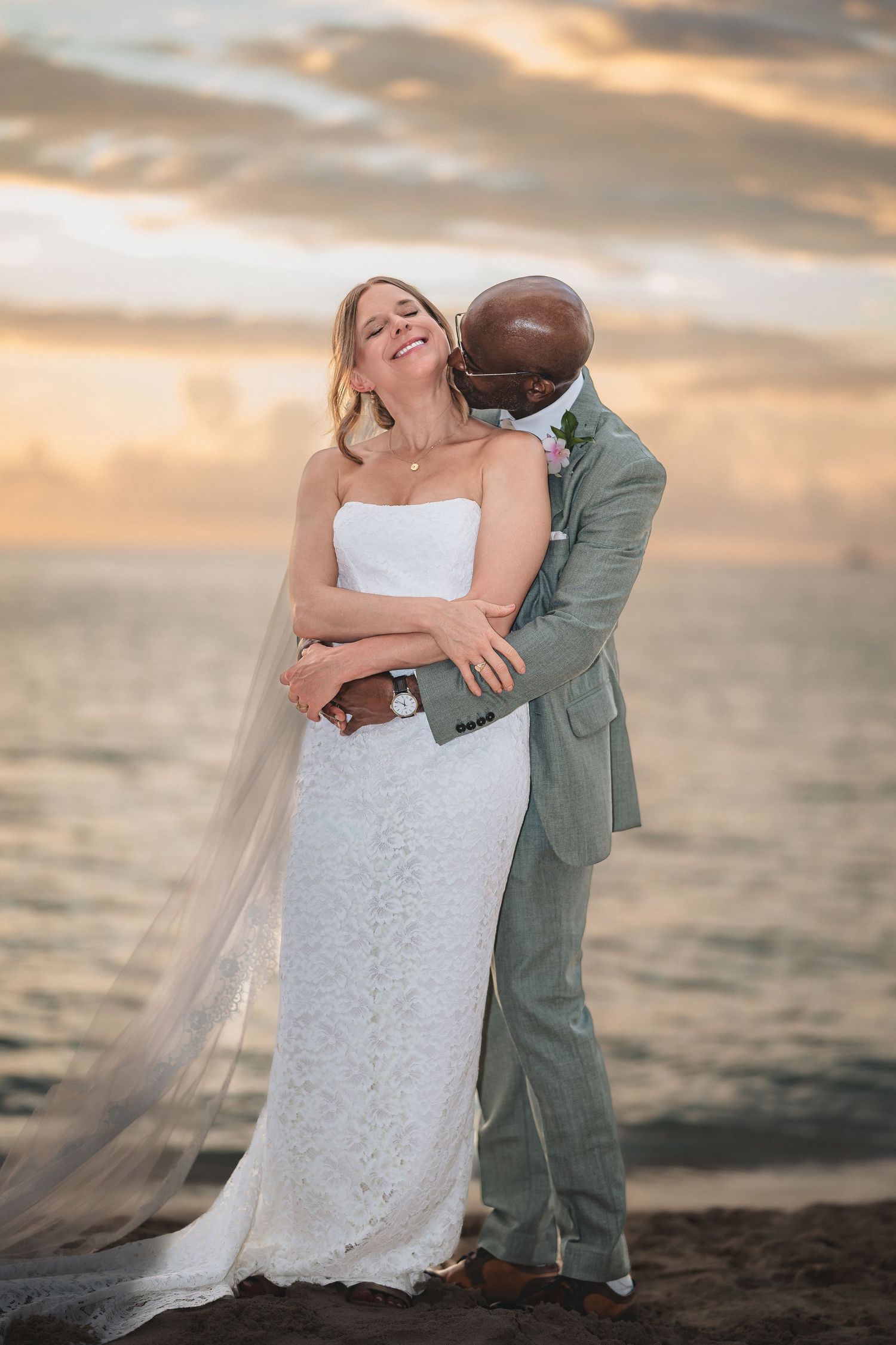 Interracial couple embracing on beach at sunset, bride in white lace gown with veil, groom in sage suit.