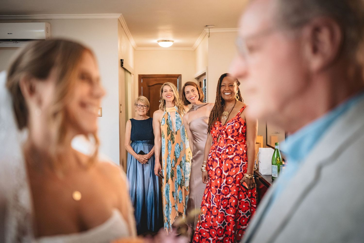 Bride smiling with bridesmaids in colorful dresses gathered in a hallway during wedding preparations.