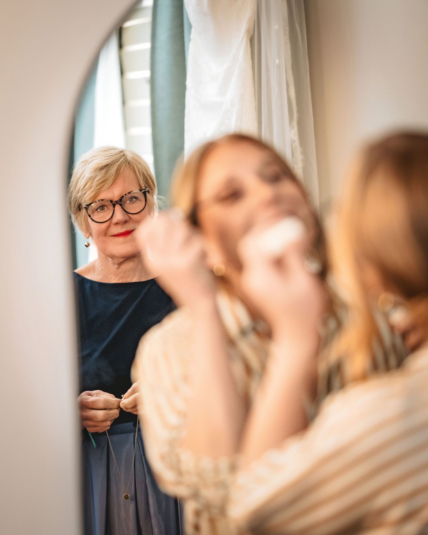 Older woman in glasses watches young blonde bride apply makeup in mirror during wedding preparations.