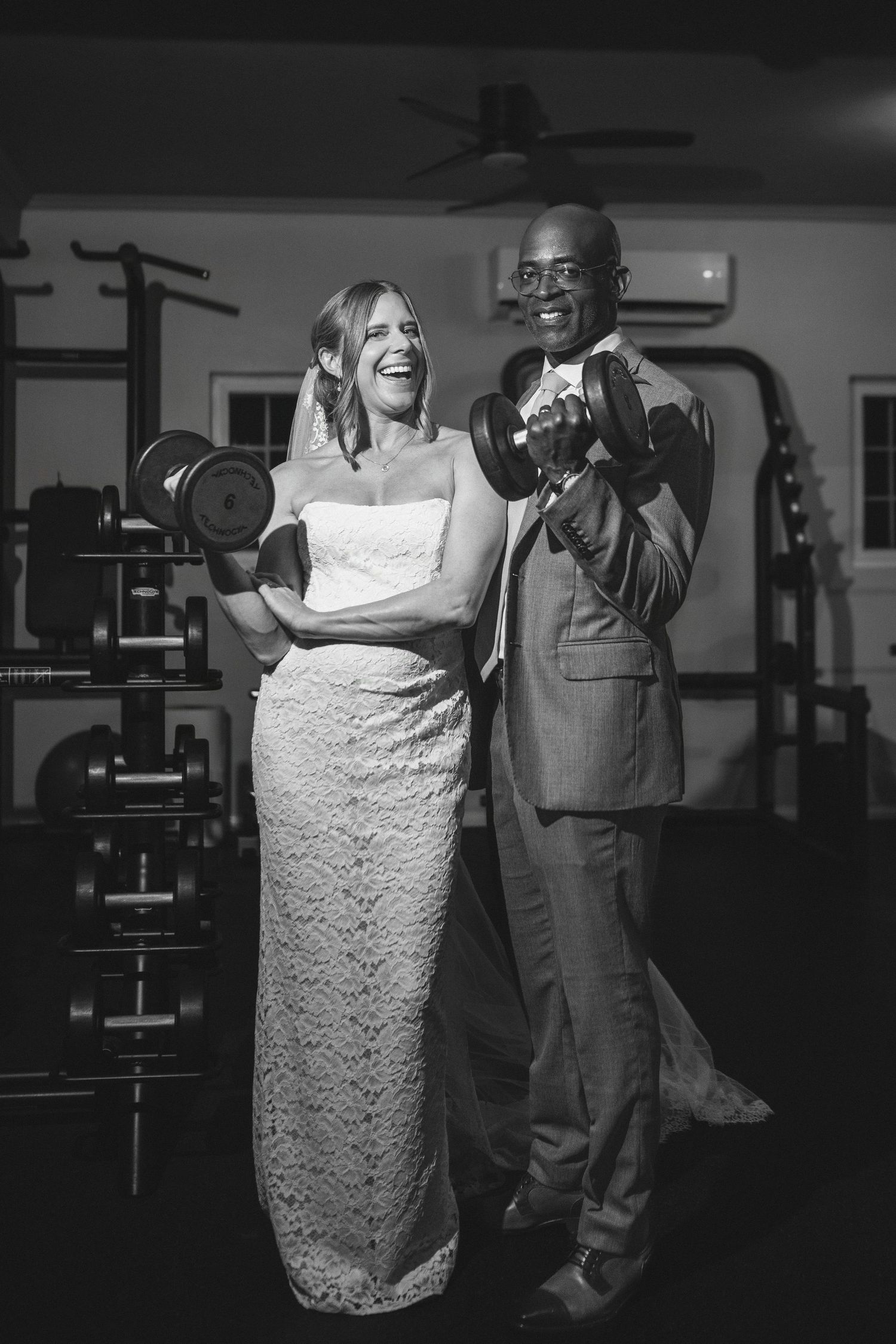 Black and white photo of a bride and groom laughing while lifting dumbbells together in a gym on their wedding day.
