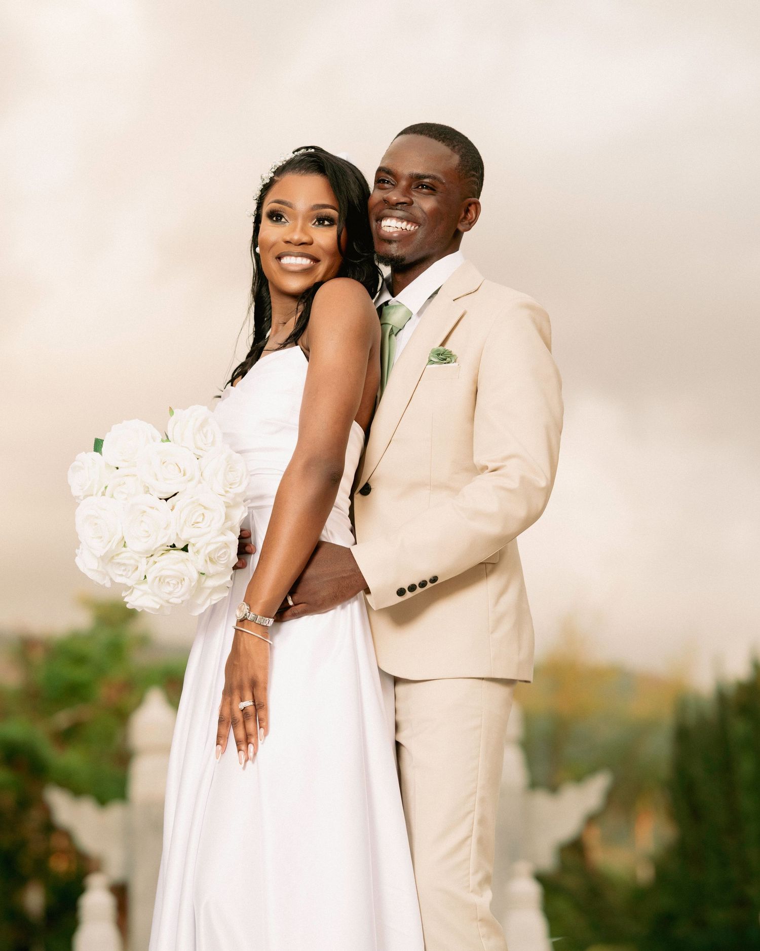 Smiling bride and groom pose together outdoors, she holds white roses bouquet, he wears beige suit with green tie.