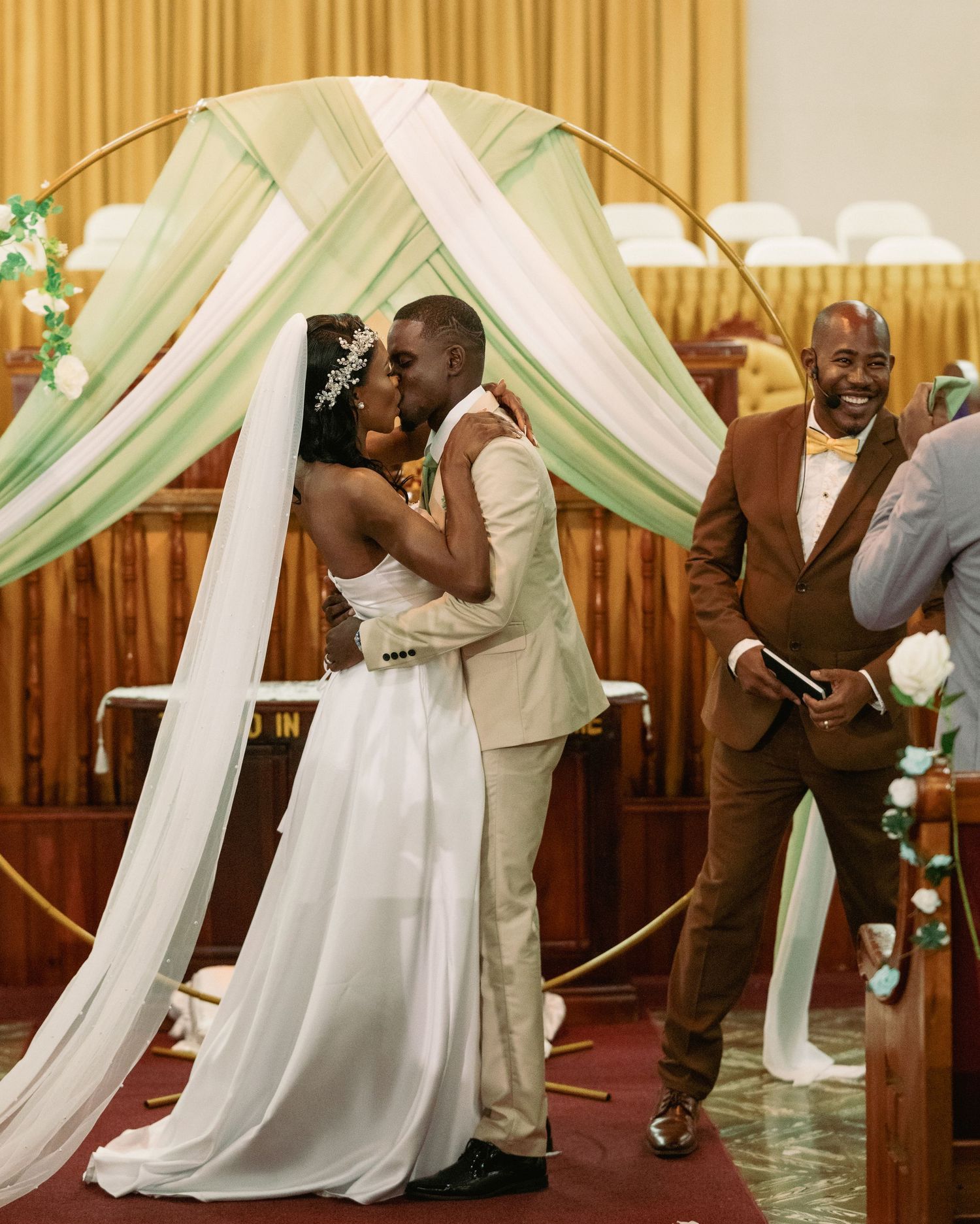 Bride and groom share first kiss at church altar under green and white draped circular arch.