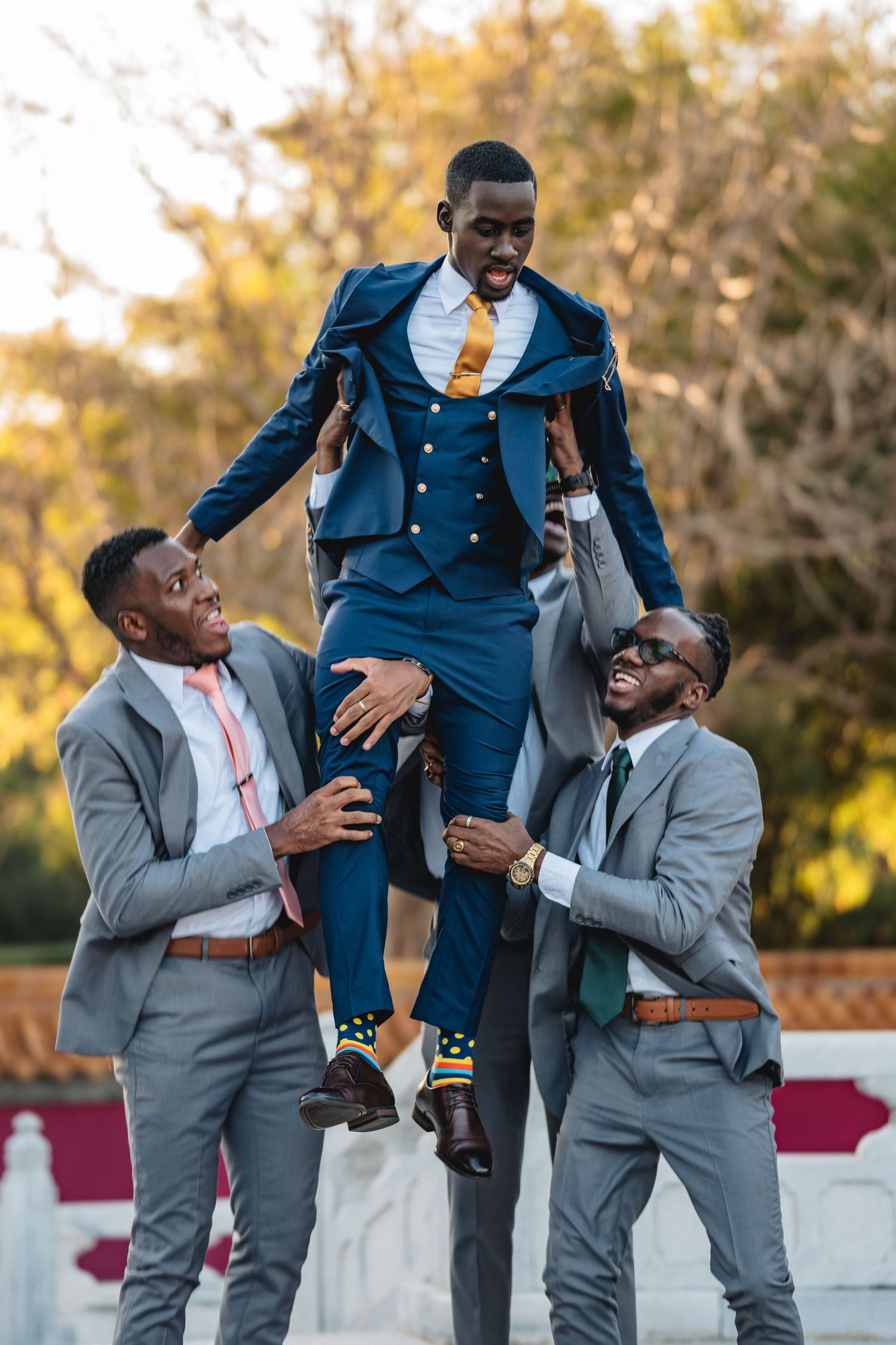 Groomsmen in gray suits joyfully lift groom in navy blue suit outdoors at wedding celebration.