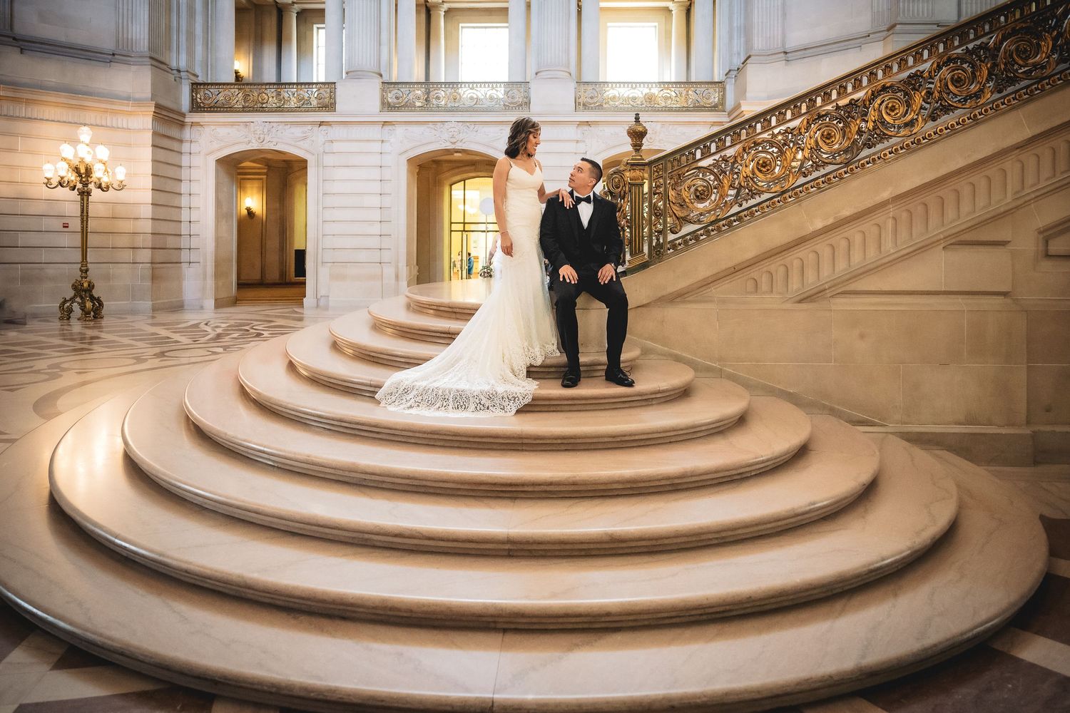 Bride and groom pose on grand staircase at San Francisco City Hall in elegant wedding attire.