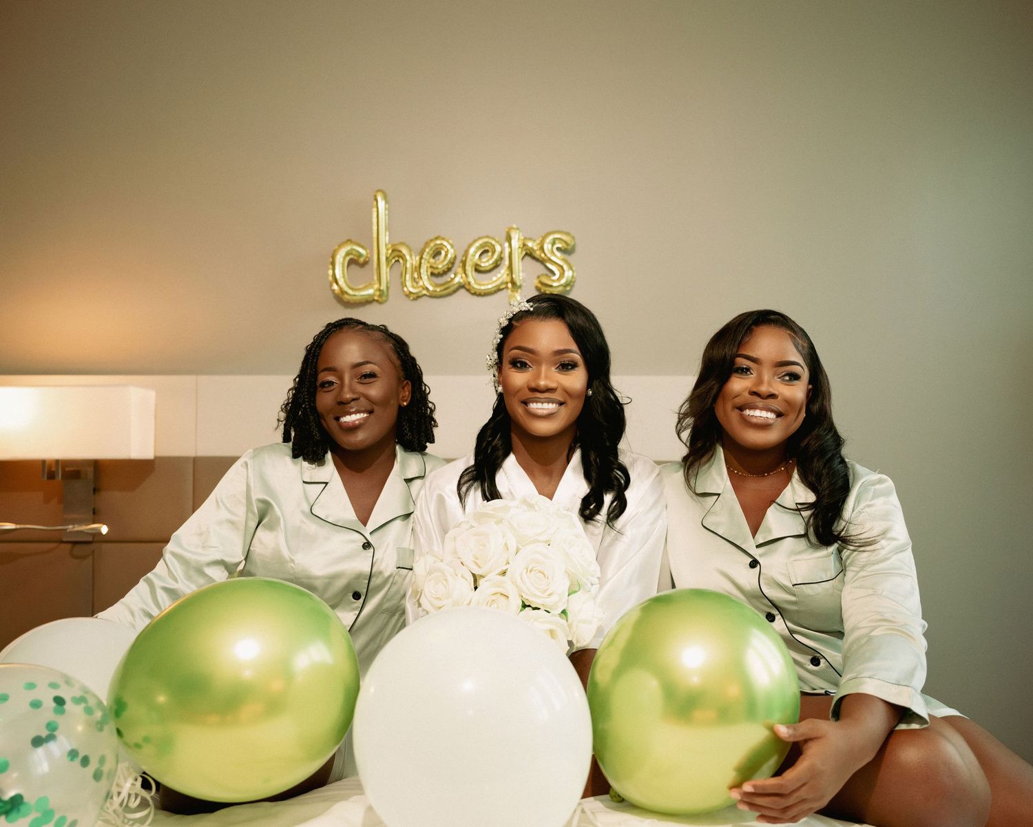 Three women in matching satin pajamas celebrating with green and white balloons under cheers sign.