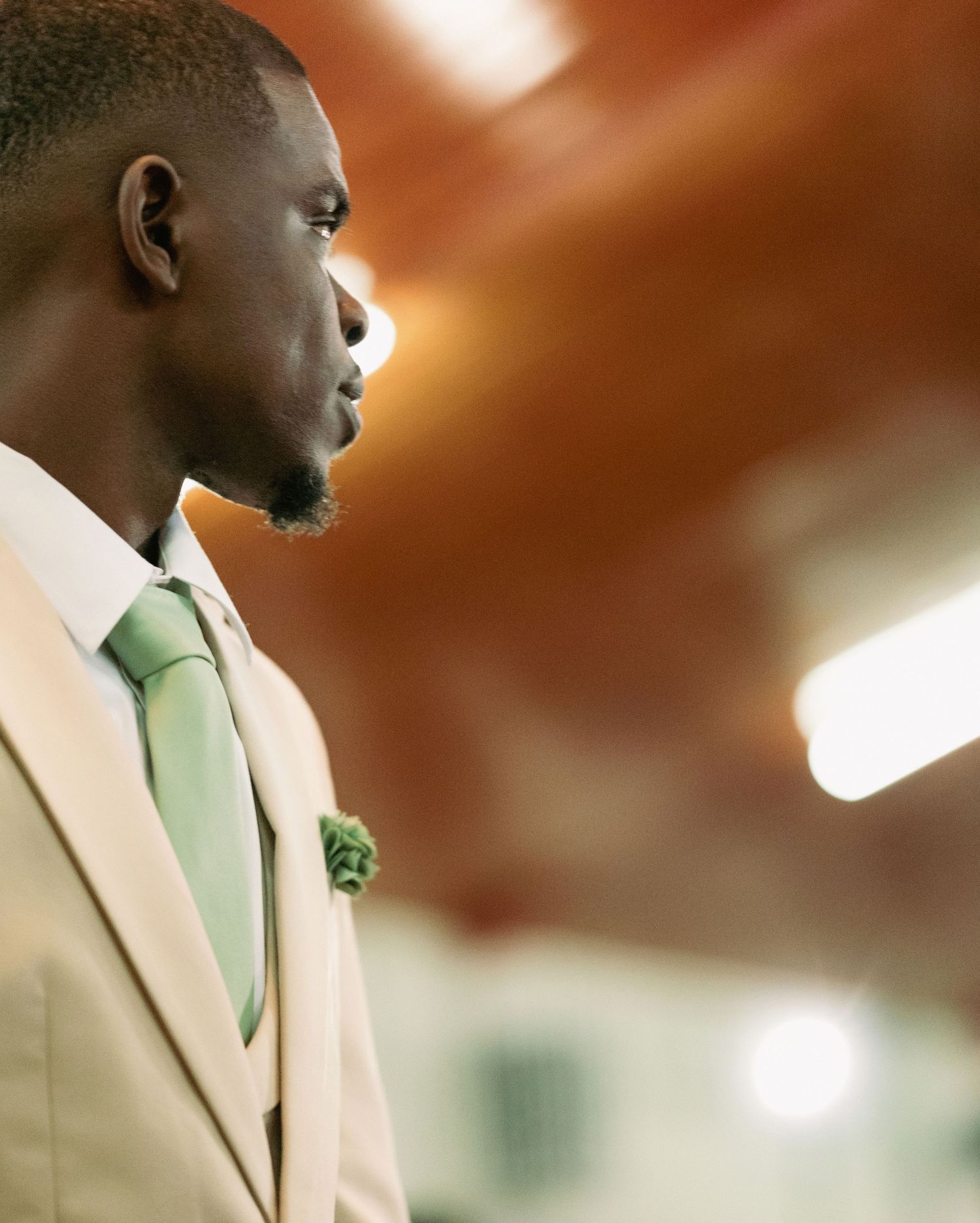Handsome Black groom in beige suit with green tie and boutonniere stands in profile view.