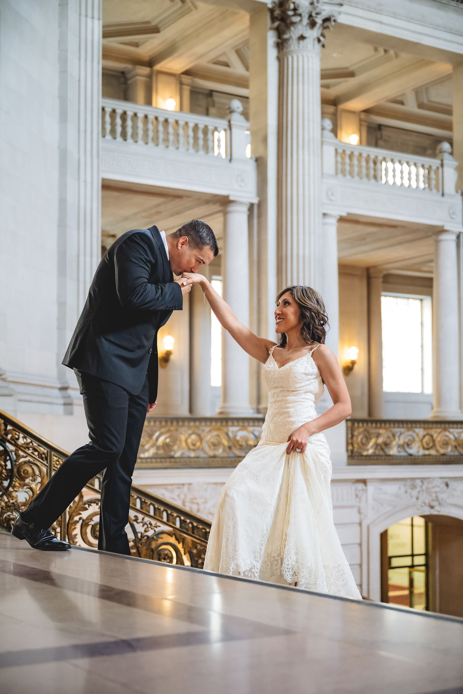 Groom kissing bride's hand on grand staircase in elegant white marble wedding venue.