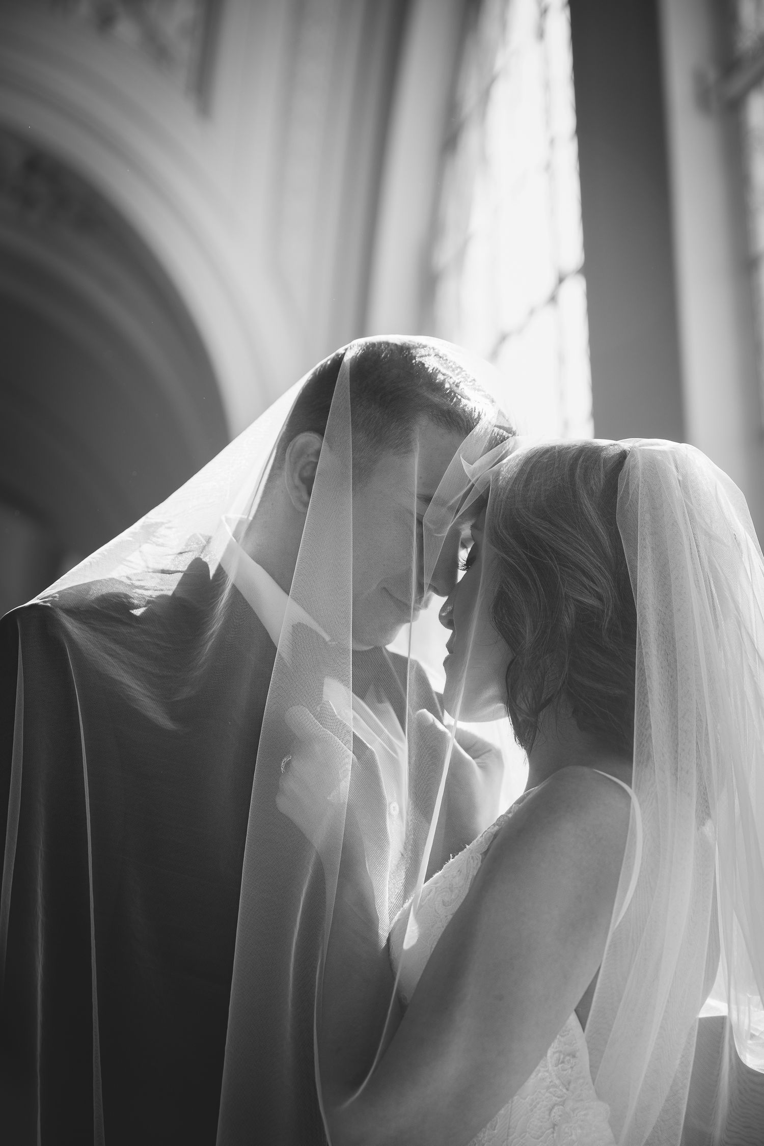Black and white photo of bride and groom sharing intimate moment under wedding veil in elegant church.